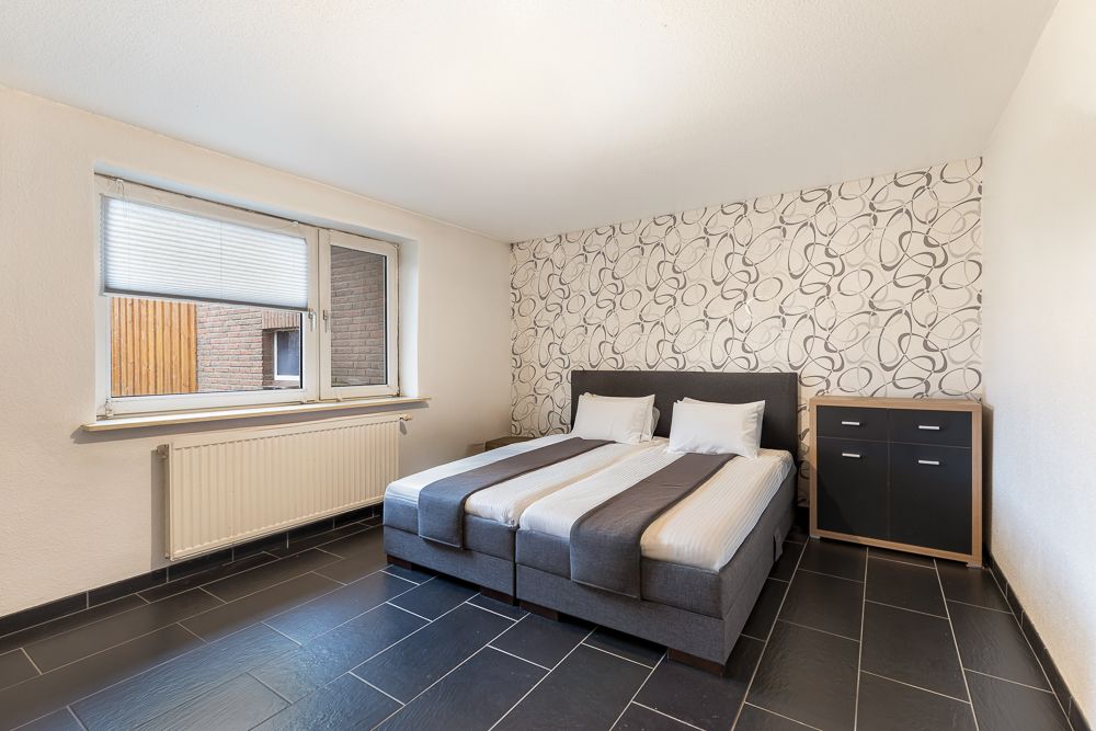 Schlafzimmer mit zwei Einzelbetten, Kommode und großem Fenster auf schwarzem Fliesenboden.