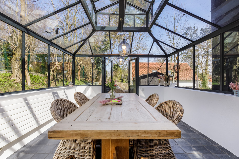 Wintergarten mit Glasdach, Holztisch und vier Korbstühlen, dekoriert mit Blumen.