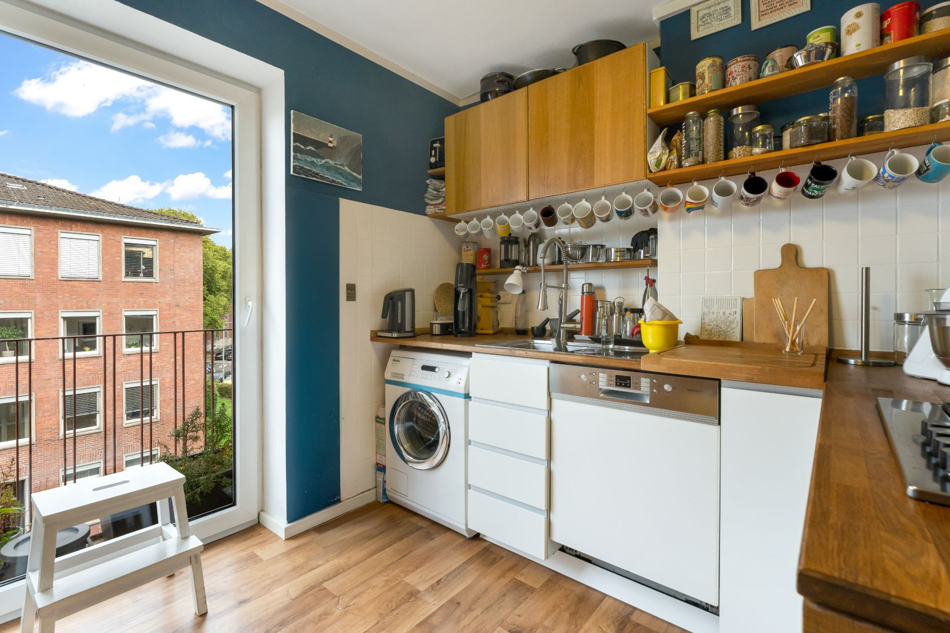 Moderne Küche mit Holzboden, weißen Schränken, offenen Regalen und großem Fenster mit Blick nach draußen.