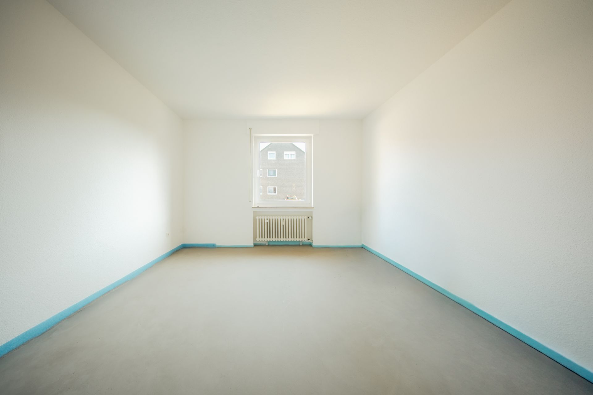 Leerer, heller Raum mit Fenster und Heizkörper, weiß gestrichene Wände, blauer Sockel.
