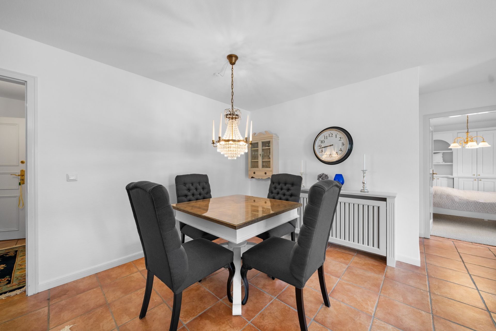Esszimmer mit vier dunklen Stühlen, Holztisch, Kronleuchter und Wanduhr auf weißer Wand.