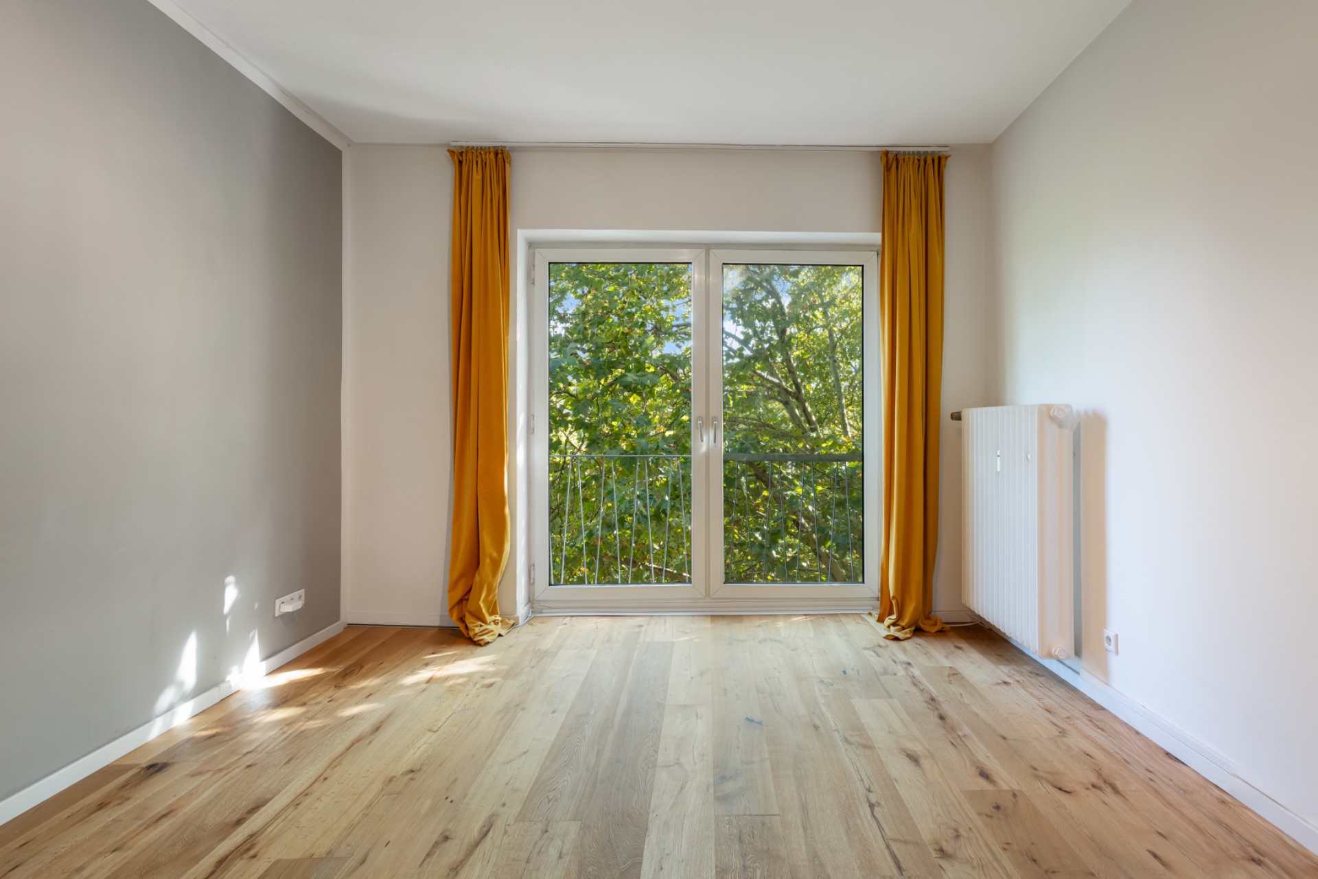 Leerer Raum mit Holzfußboden, großen Fenstern und gelben Vorhängen, Blick auf grüne Bäume.