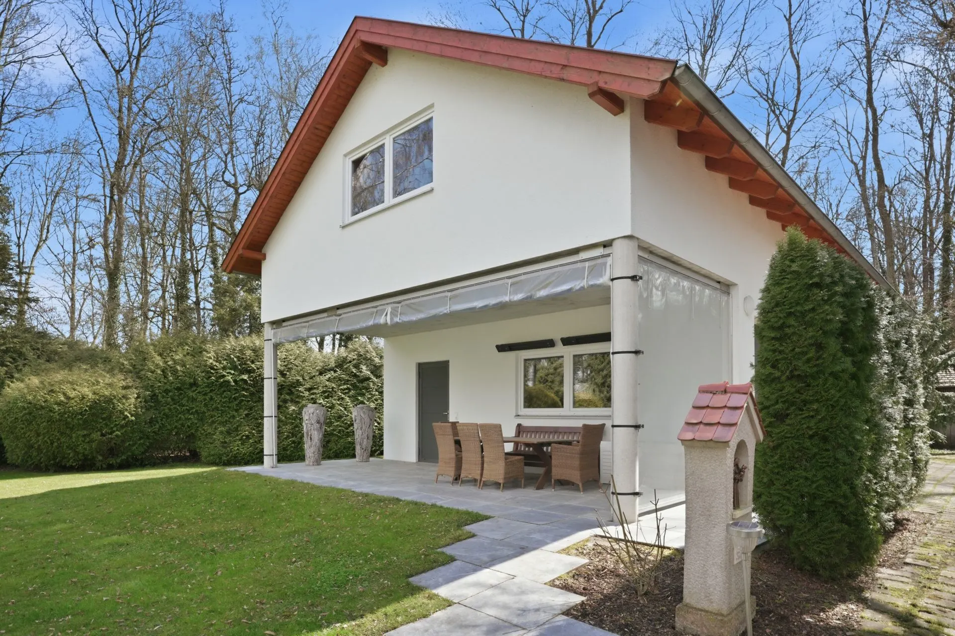 Modernes weißes Haus mit Terrasse, Esstisch und Garten, umgeben von Bäumen.