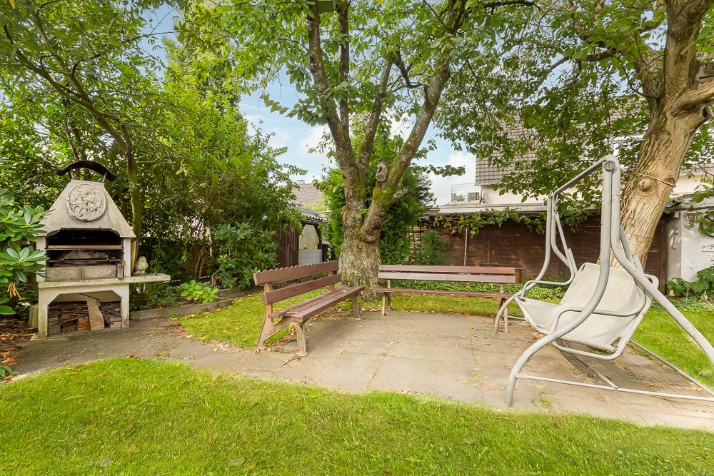 Garten mit Steingrill, zwei Holzbänken und einer Schaukelbank unter Bäumen.