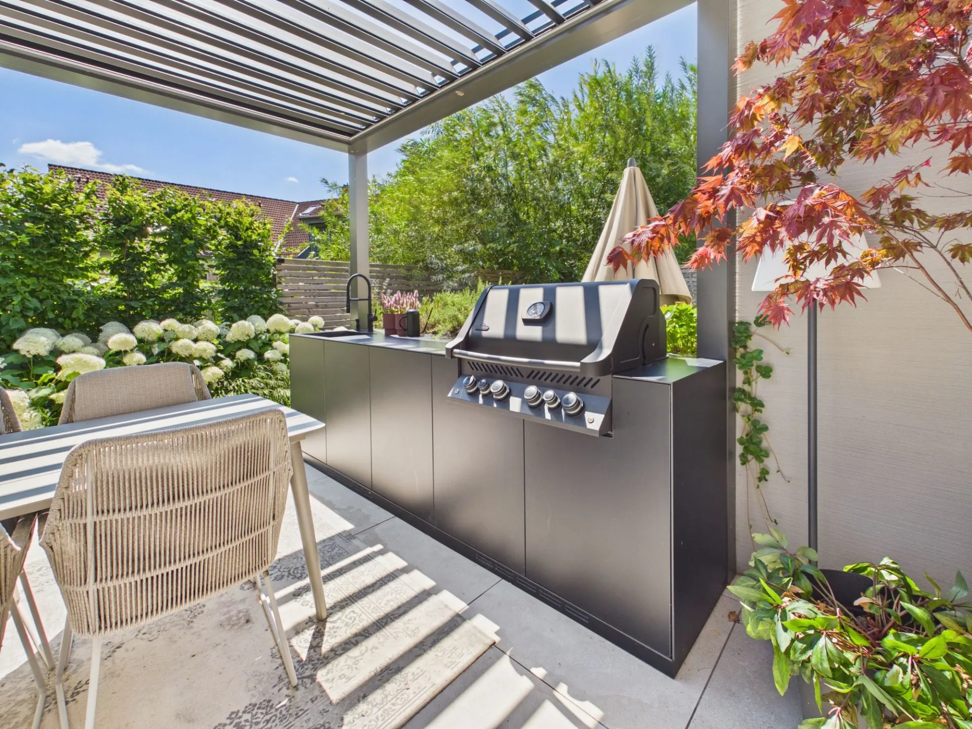 Moderne Outdoorküche mit Grill und Spüle auf einer überdachten Terrasse, umgeben von Pflanzen.