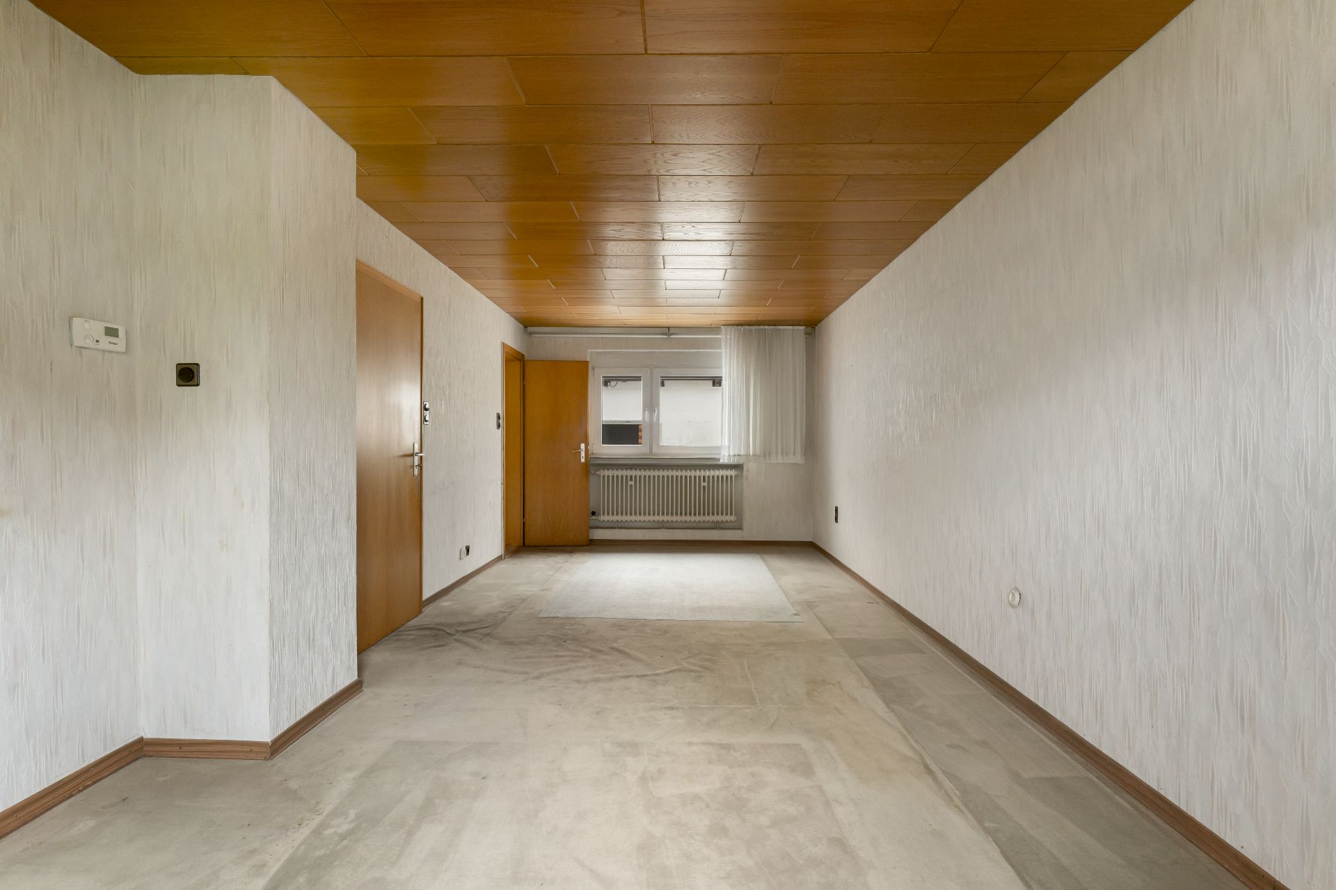 Leerer Raum mit Holzdecke, zwei Türen, Fenster und Heizkörper an der Stirnseite.