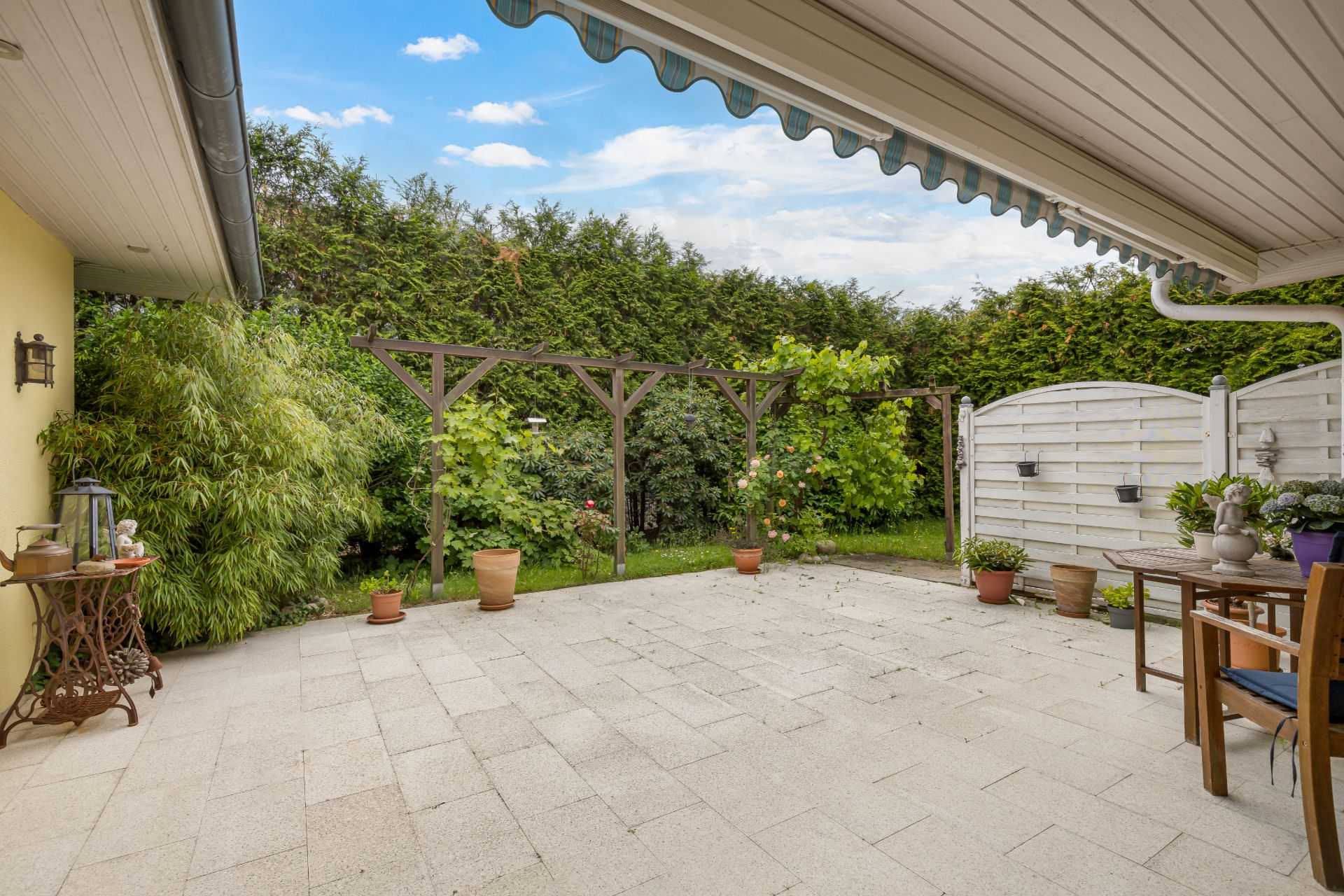 Gepflasterte Terrasse mit Tisch, Stühlen, Pflanzen und Markise, begrenzt von Hecken und Zaun.