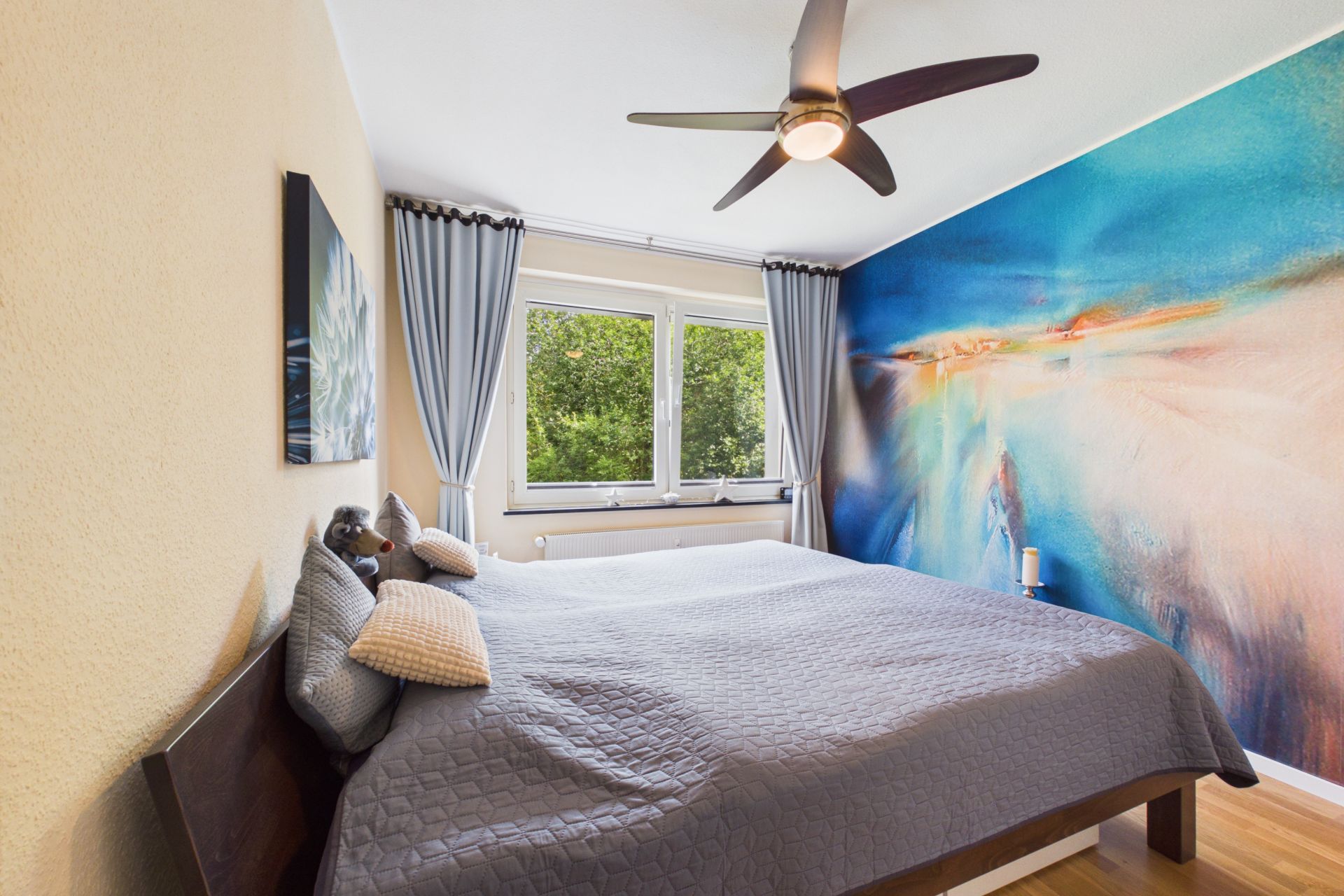 Schlafzimmer mit großem Bett, bunter Wand, Deckenventilator und Fenstern mit Blick ins Grüne.