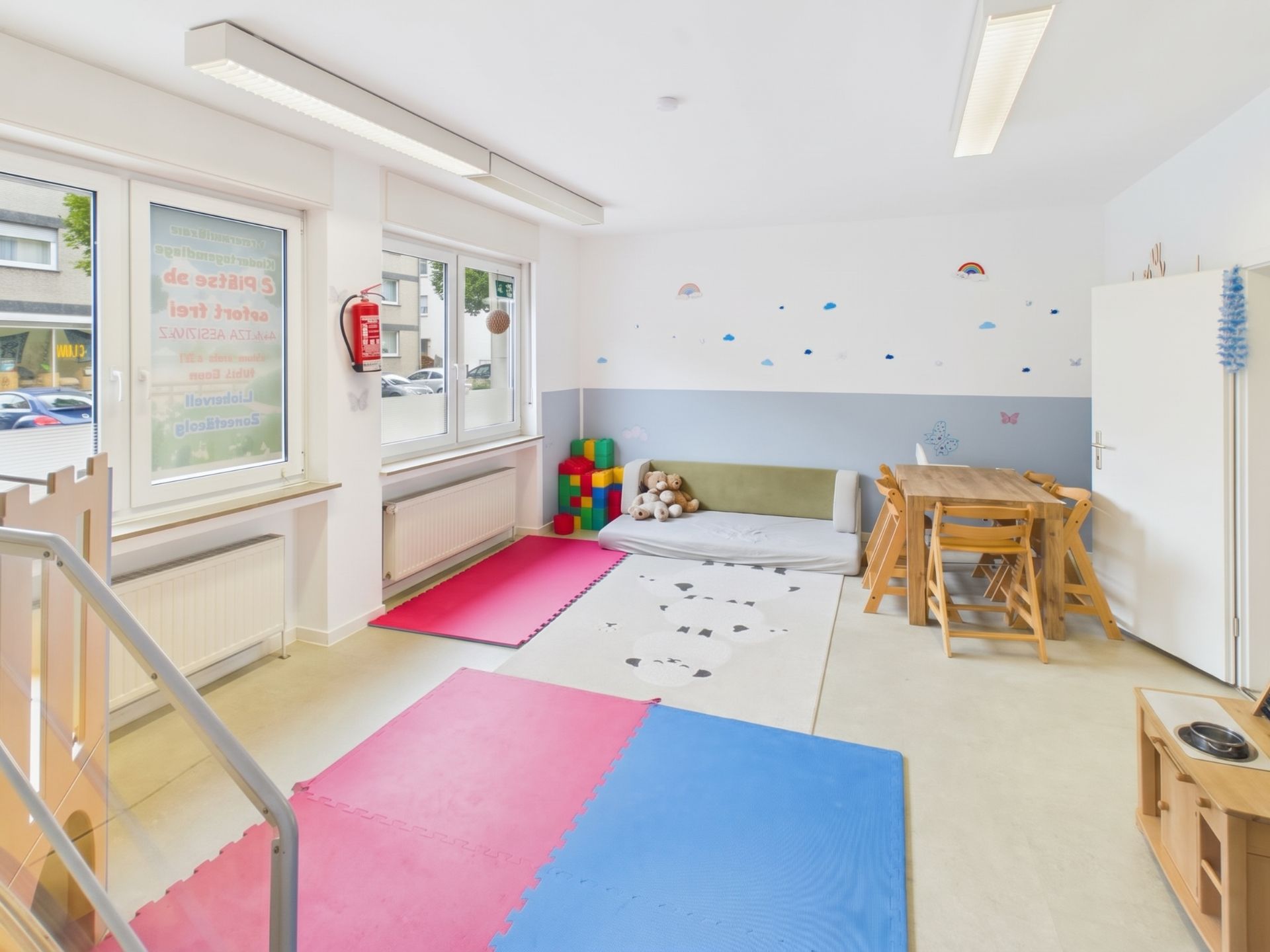 Heller Raum mit bunten Spielmatten, Sitzbereich mit Kuscheltieren und einem Tisch mit Stühlen.