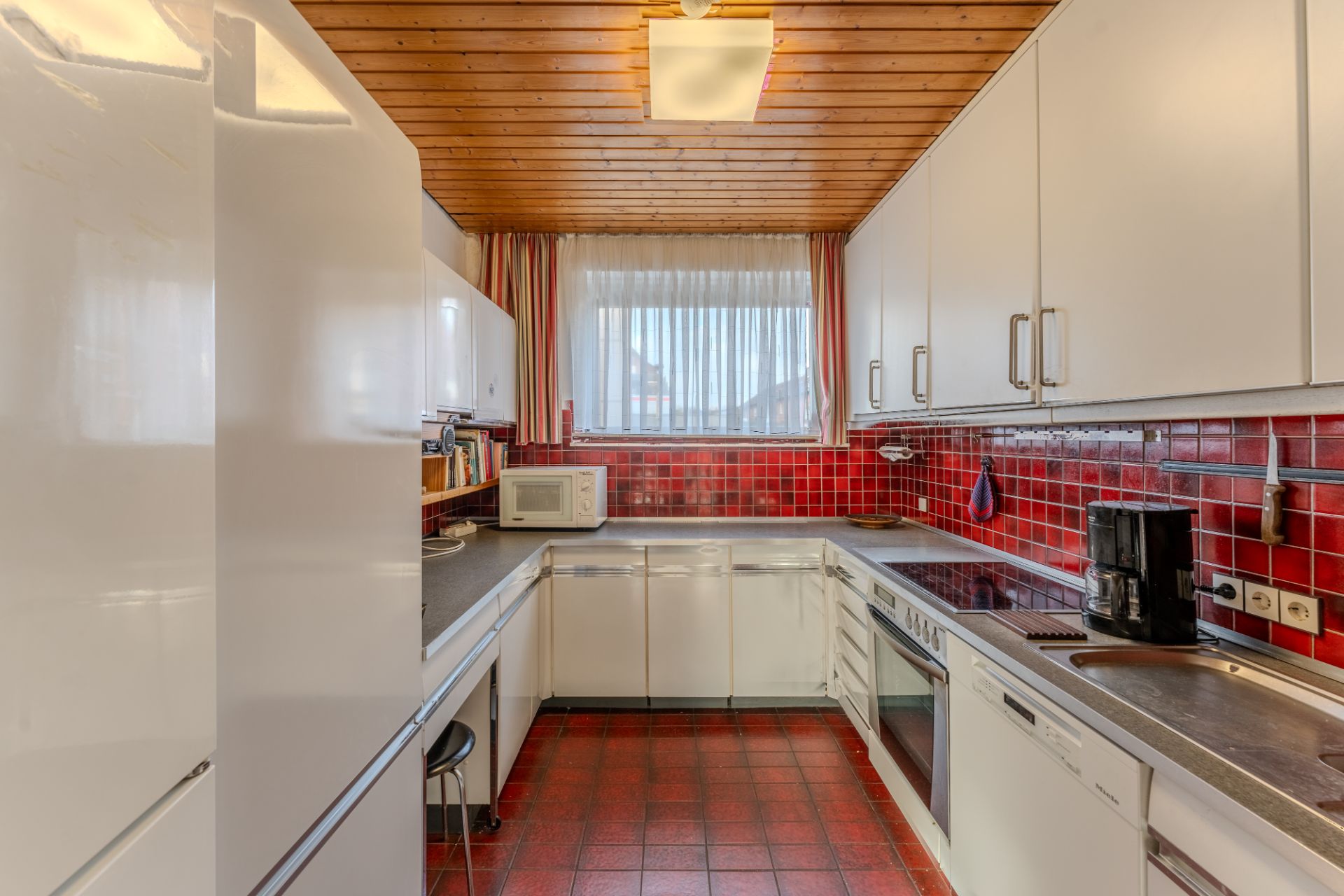 Küche mit weißen Schränken, roten Fliesen, Holzbalkendecke und großem Fenster.