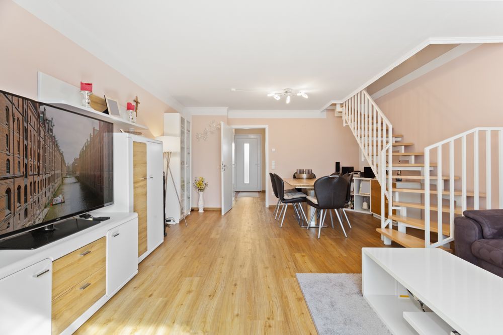 Modernes Wohnzimmer mit Holzfußboden, TV, Esstisch, Regalwand und offener Treppe nach oben.