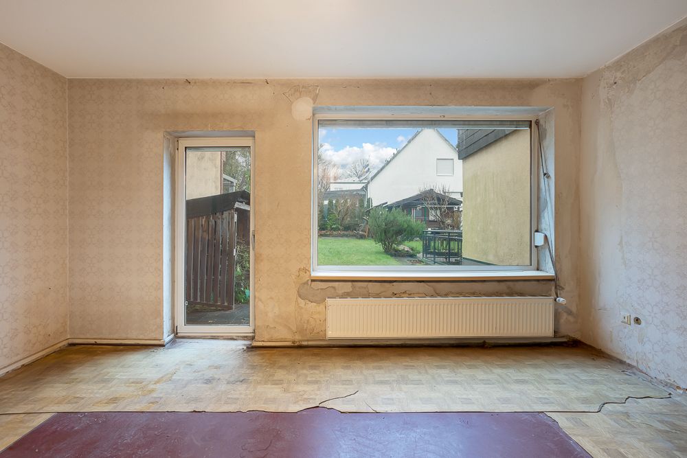 Leeres Zimmer mit großen Fenstern und Tür zum Garten, abgenutzte Wände und Boden.