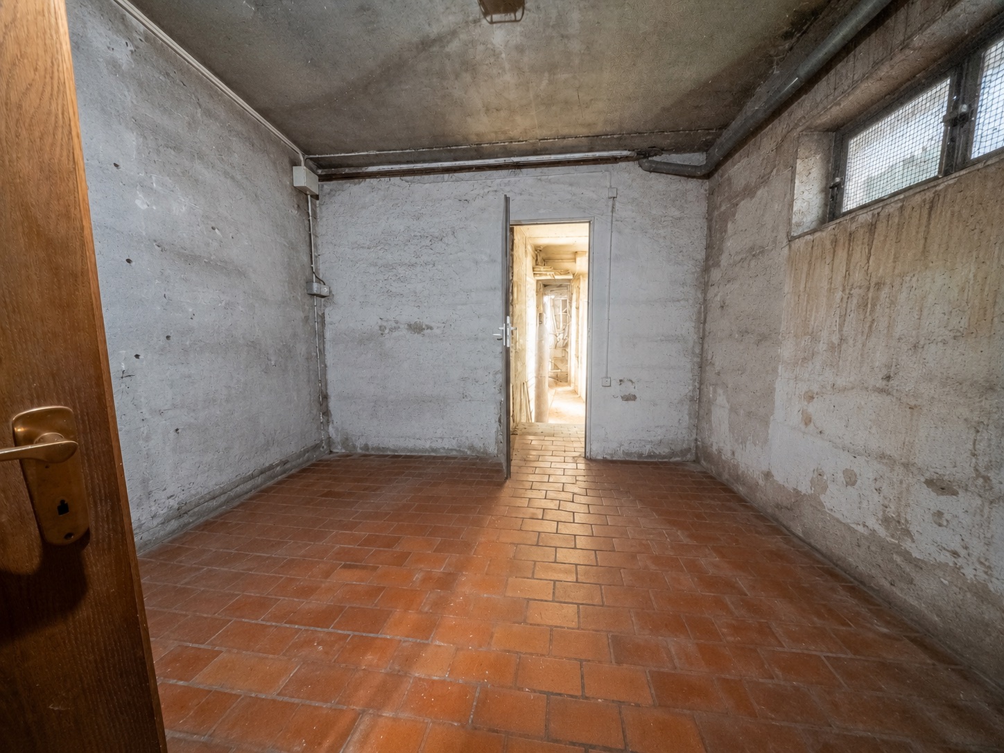 Leerer Kellerraum mit Fliesenboden, Betonwänden und kleinem Fenster.