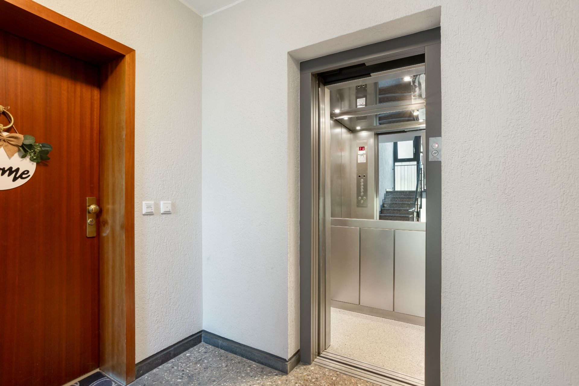 Geöffneter Aufzug neben einer Wohnungstür mit Dekoration im Flur eines Wohngebäudes.