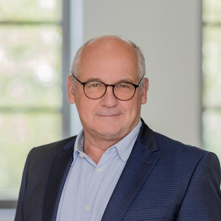 Ein älterer Mann mit Brille und blauer Jacke steht vor einem Fenster.