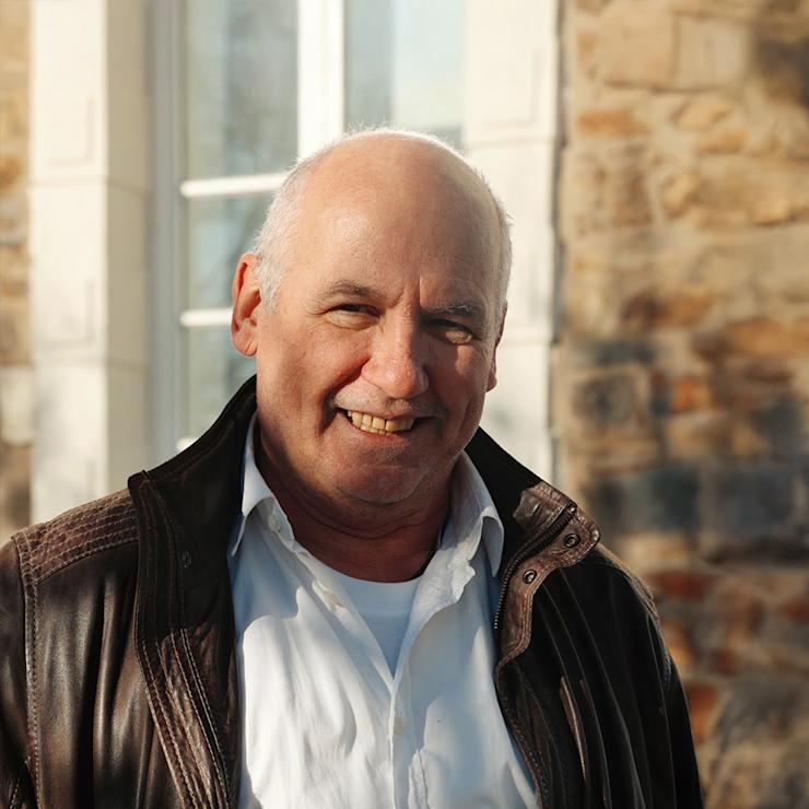 Ein älterer Mann mit Brille und blauer Jacke steht vor einem Fenster.