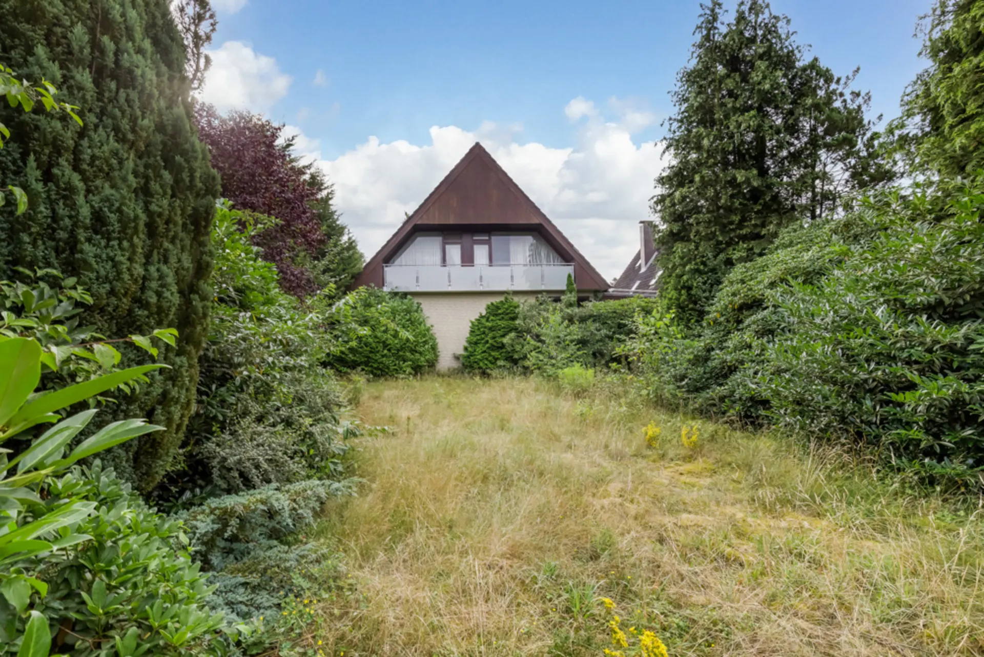 Haus mit spitzem Dach und Balkon, umgeben von dicht bewachsenem Garten mit hohem Gras.