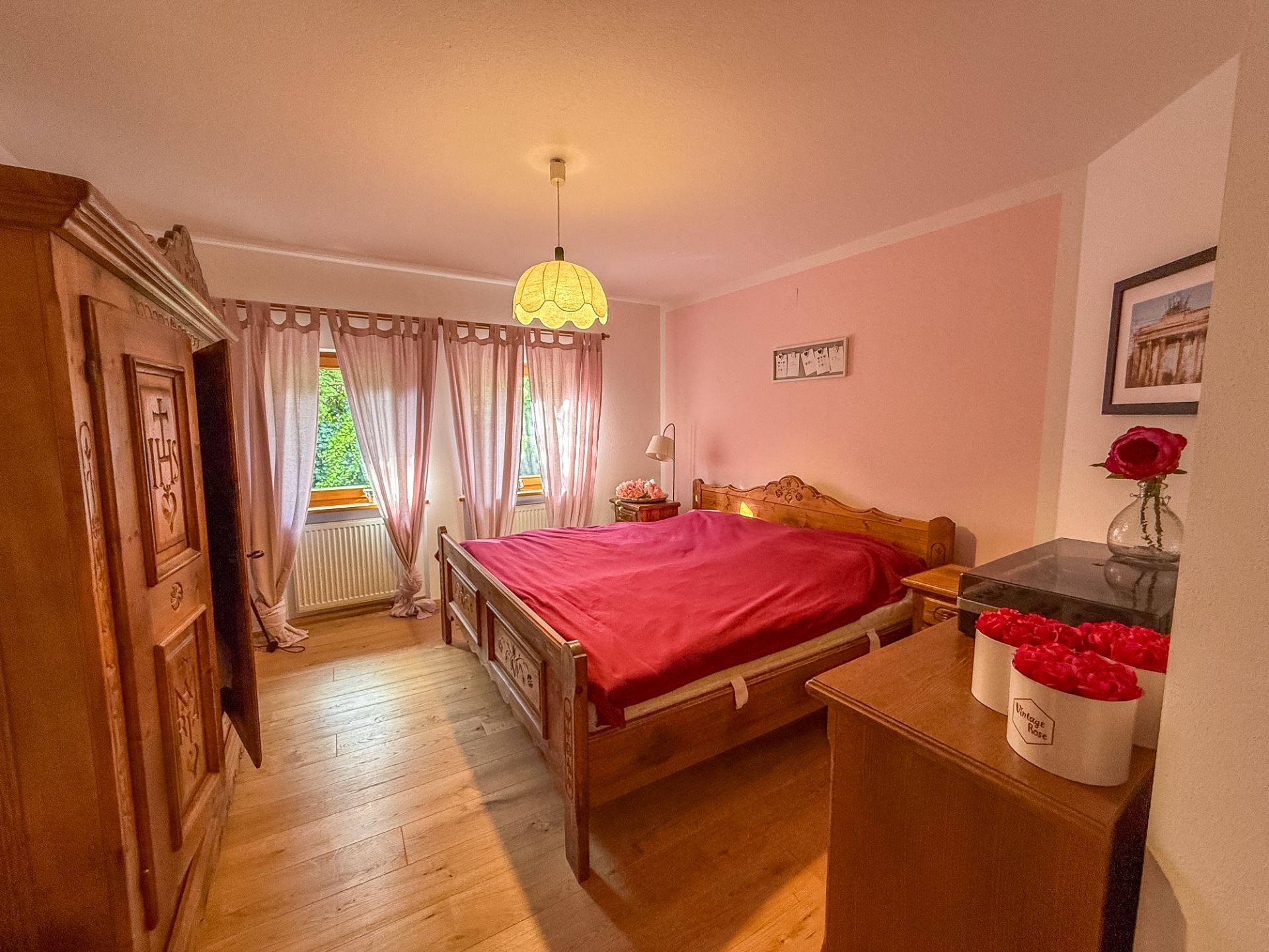 Schlafzimmer mit Holzbett, roter Bettdecke, Kommode und großen Fenstern mit rosa Vorhängen.