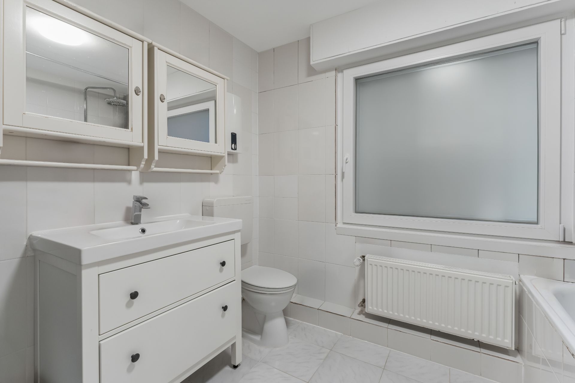 Helles Badezimmer mit Waschbecken, Spiegel, Toilette und großem Fenster mit Milchglas.