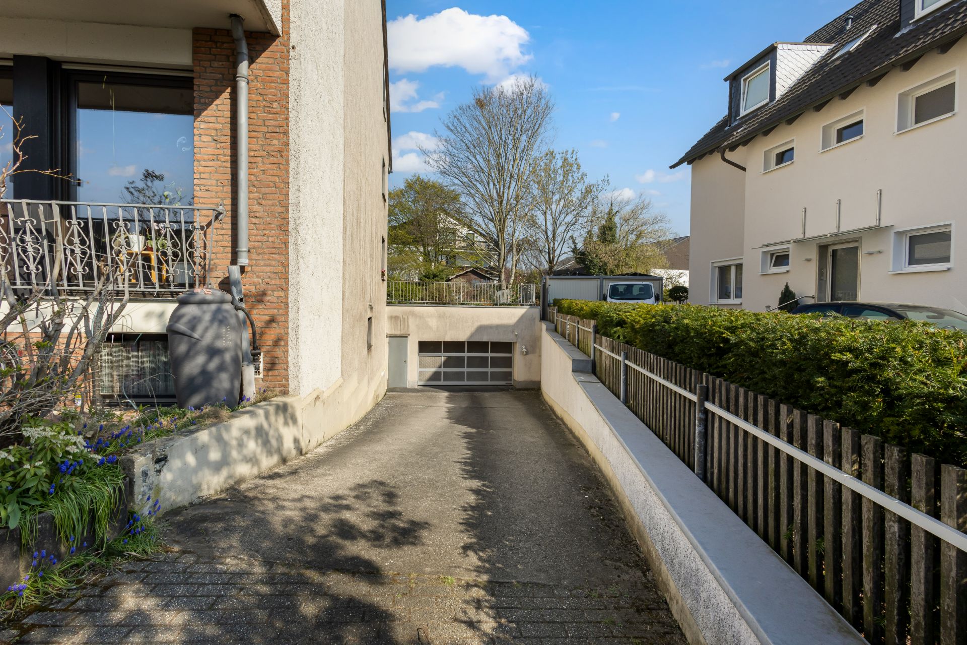 Auffahrt zu einer Garage unter einem Wohnhaus mit Zaun und Pflanzen am Rand.