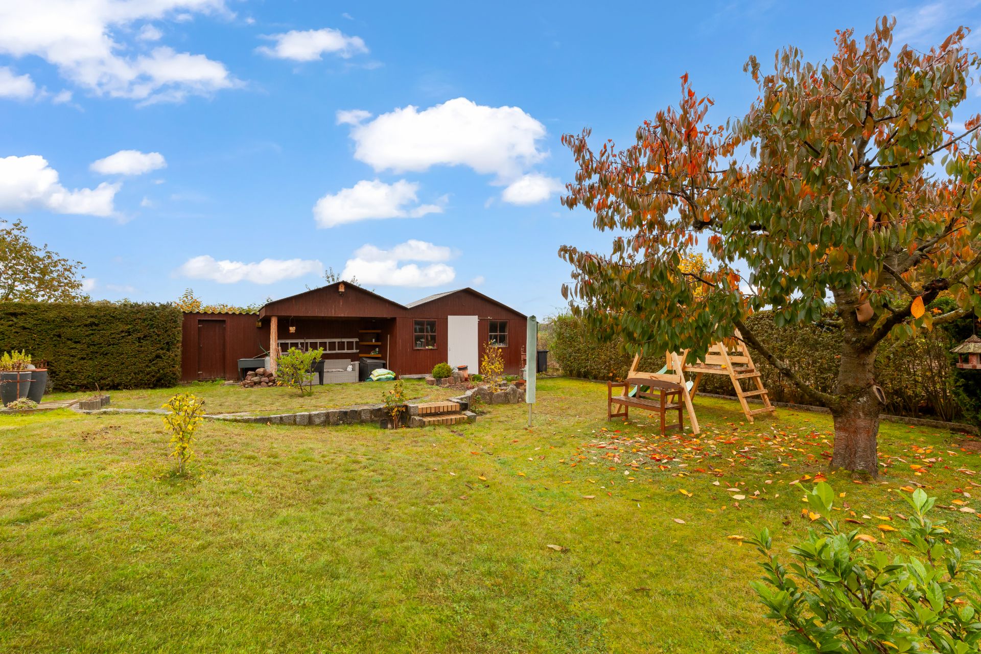 Garten mit grünem Rasen, Holzhütte, Baum und Sitzbank unter blauem Himmel.