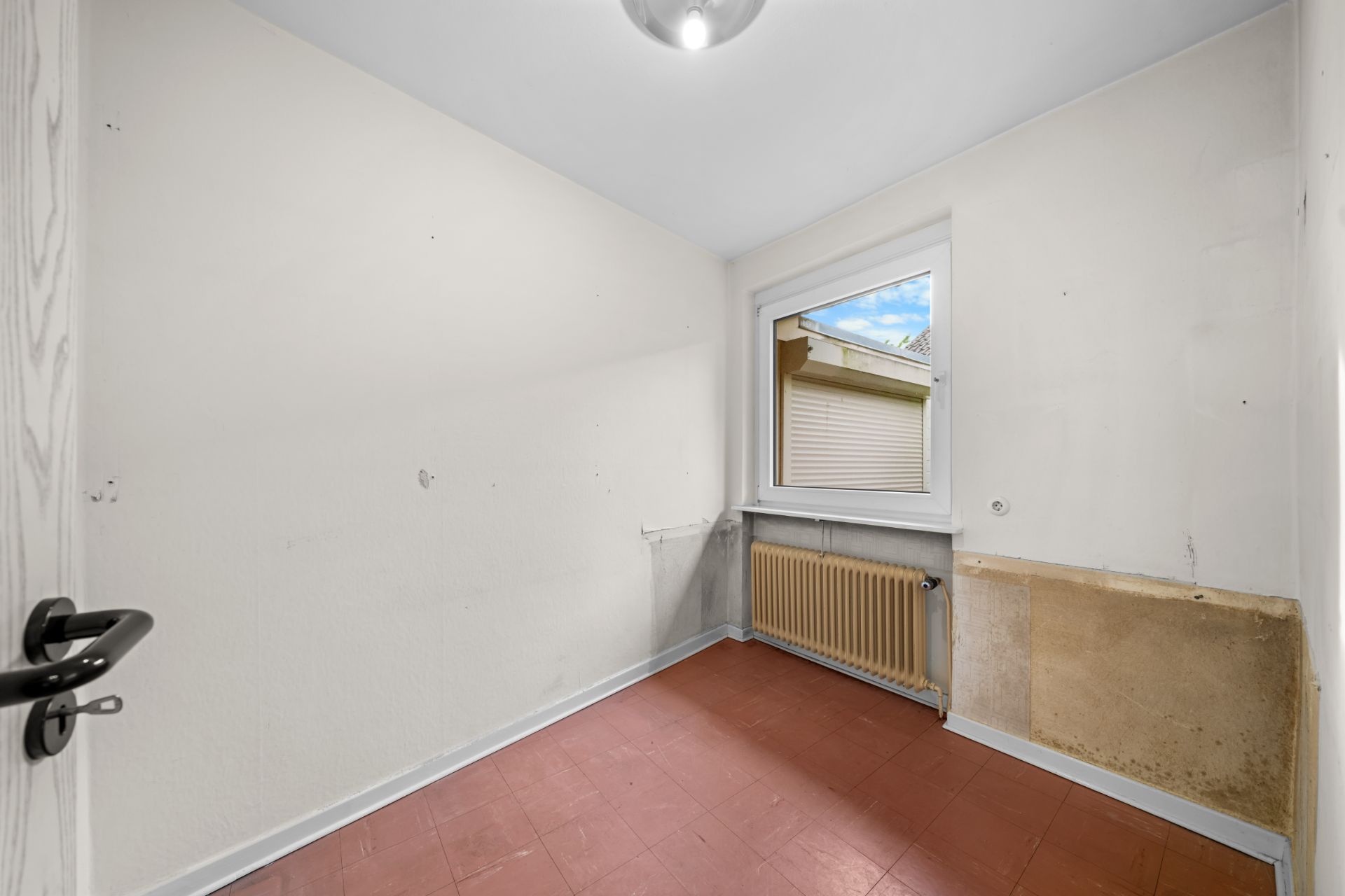 Kleines, leerstehendes Zimmer mit Fenster, Heizkörper und roten Bodenfliesen.