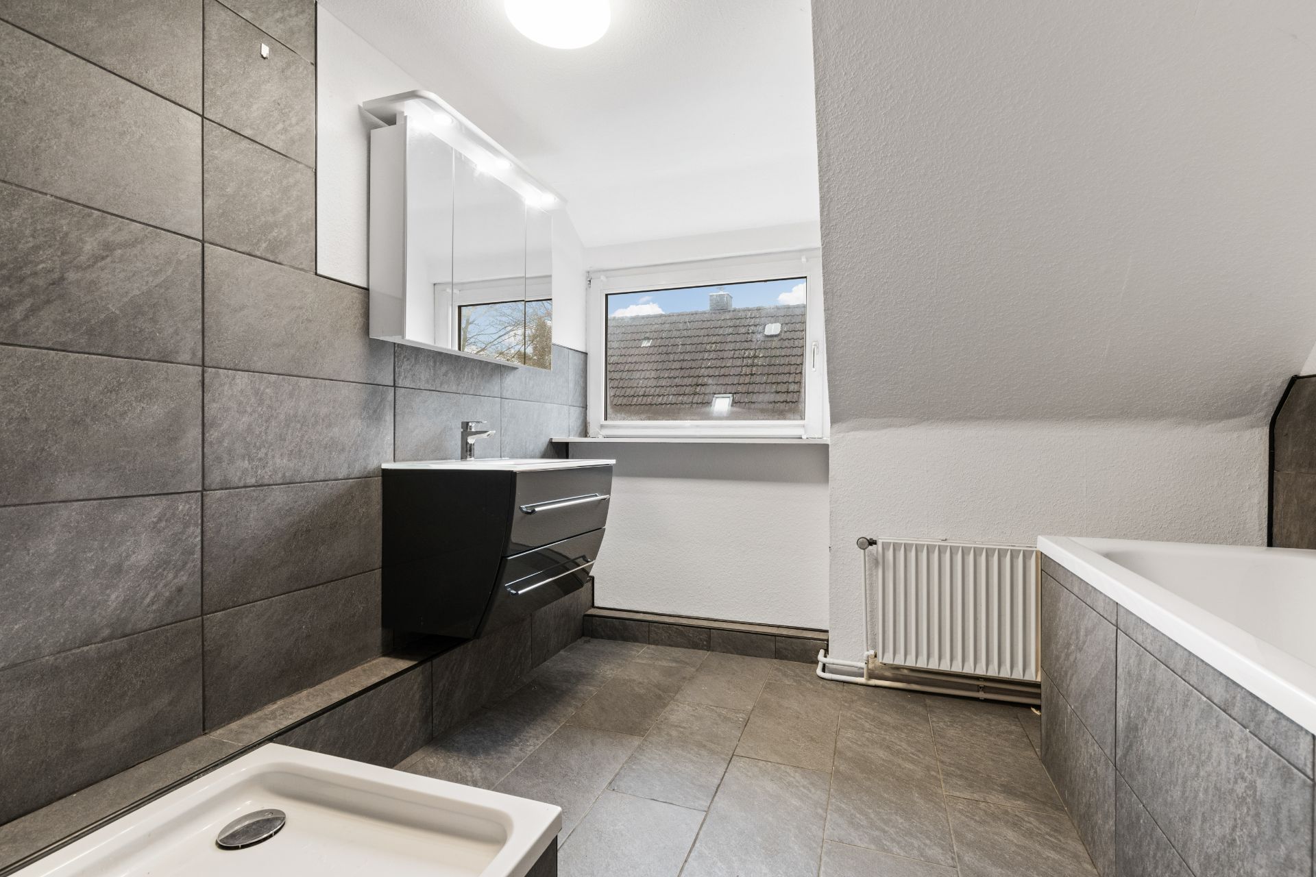 Modernes Badezimmer mit grauen Fliesen, Badewanne, Waschbecken und großem Fenster.