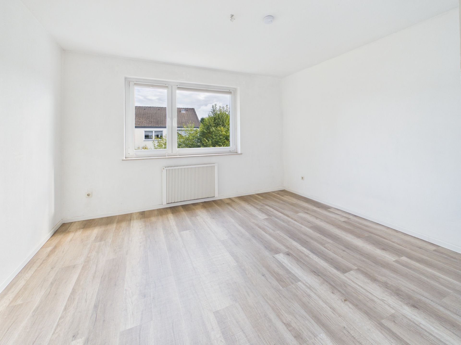 Leerer, heller Raum mit großem Fenster, weißer Wand und hellem Holzfußboden.