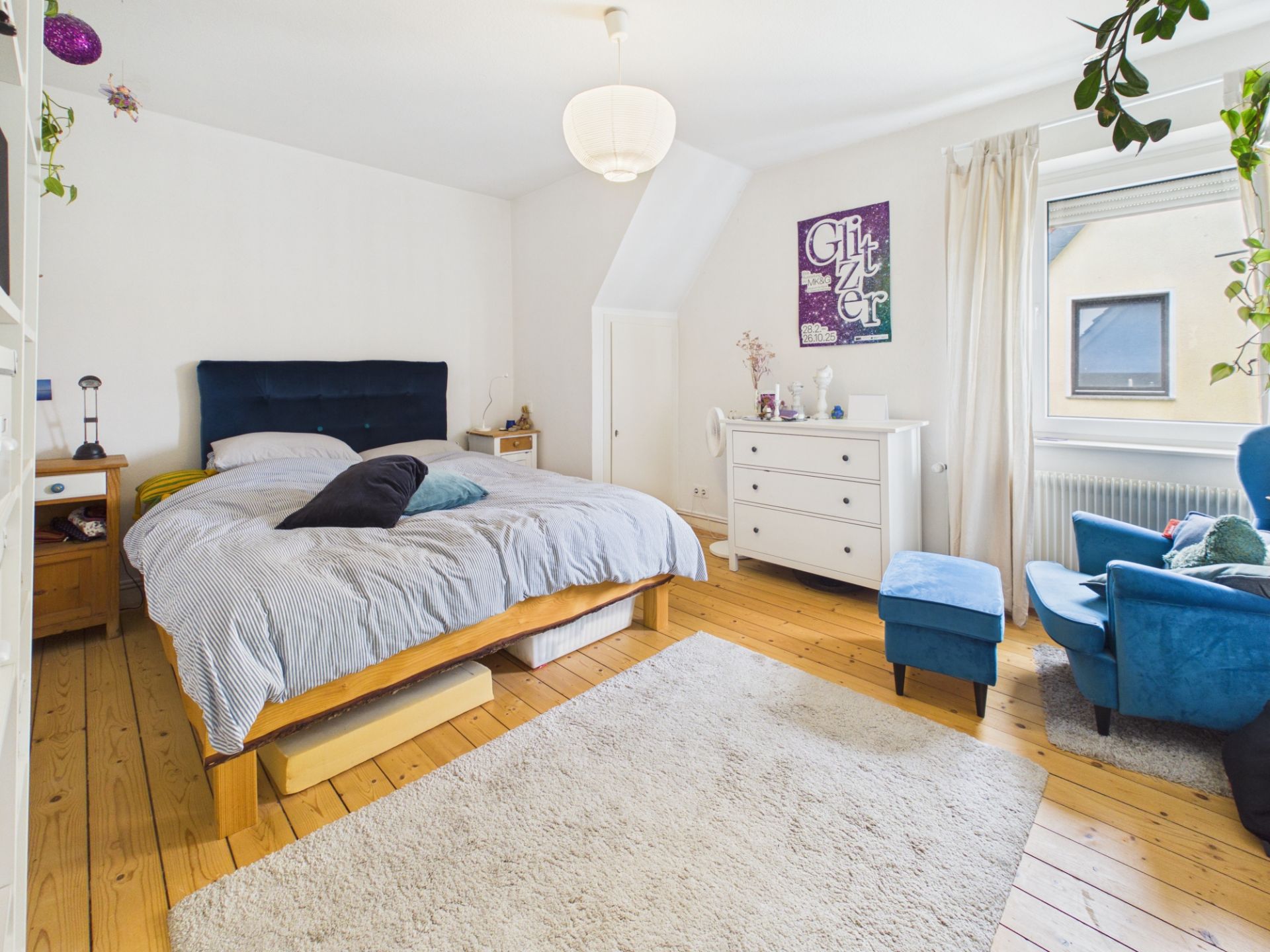 Helles Schlafzimmer mit Doppelbett, Teppich, blauer Sessel, Kommode und Fenster.