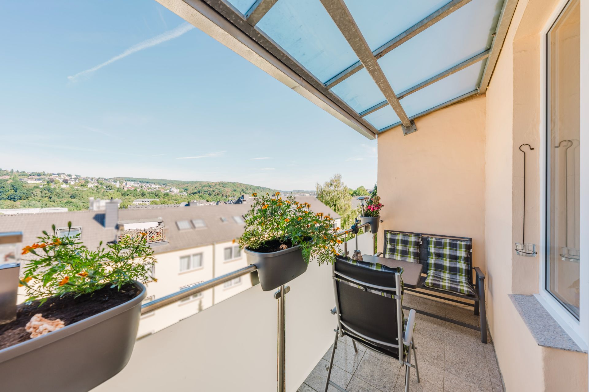 Überdachter Balkon mit Sitzmöbeln und Blumenkästen, Blick auf Nachbarhäuser und grüne Hügel.