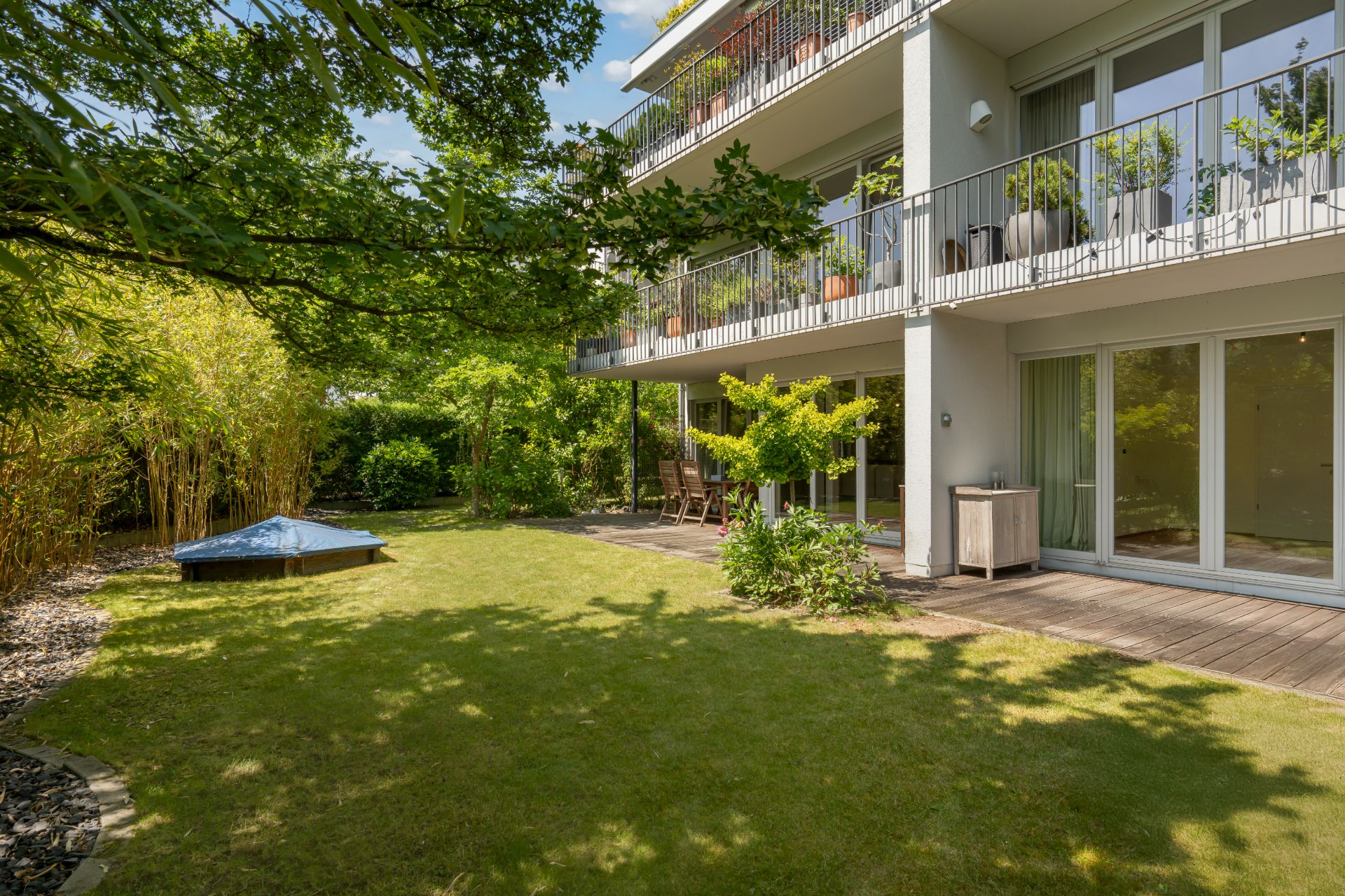 Grüner Garten mit Terrasse und Balkon an einem modernen Wohngebäude, umgeben von Bäumen und Sträuchern.