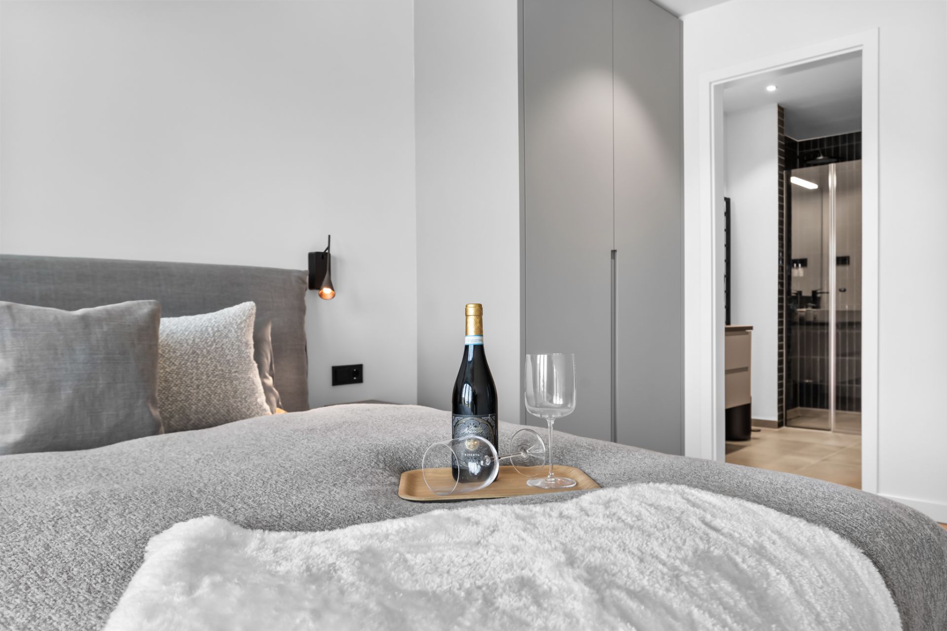 Modernes Schlafzimmer mit grauem Bett, Tablett, Weinflasche und Gläsern, Blick ins Badezimmer.