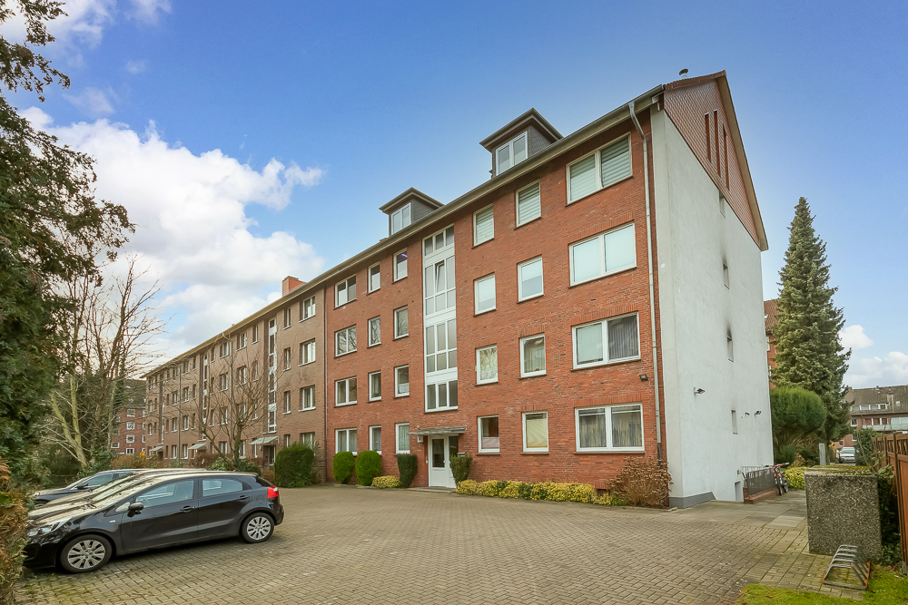 Mehrfamilienhaus aus rotem Backstein mit mehreren Fenstern und parkenden Autos im Vordergrund.
