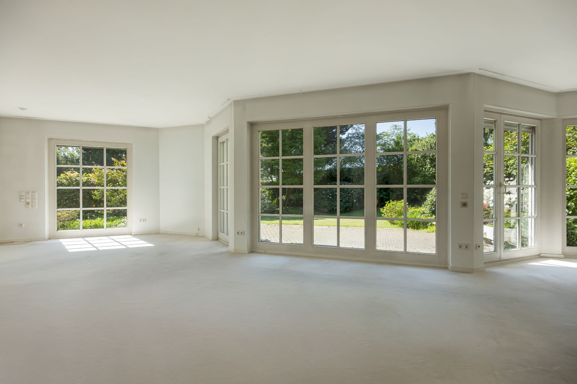 Großer, leerer Raum mit weißen Wänden und großen Fenstern mit Blick auf einen Garten.