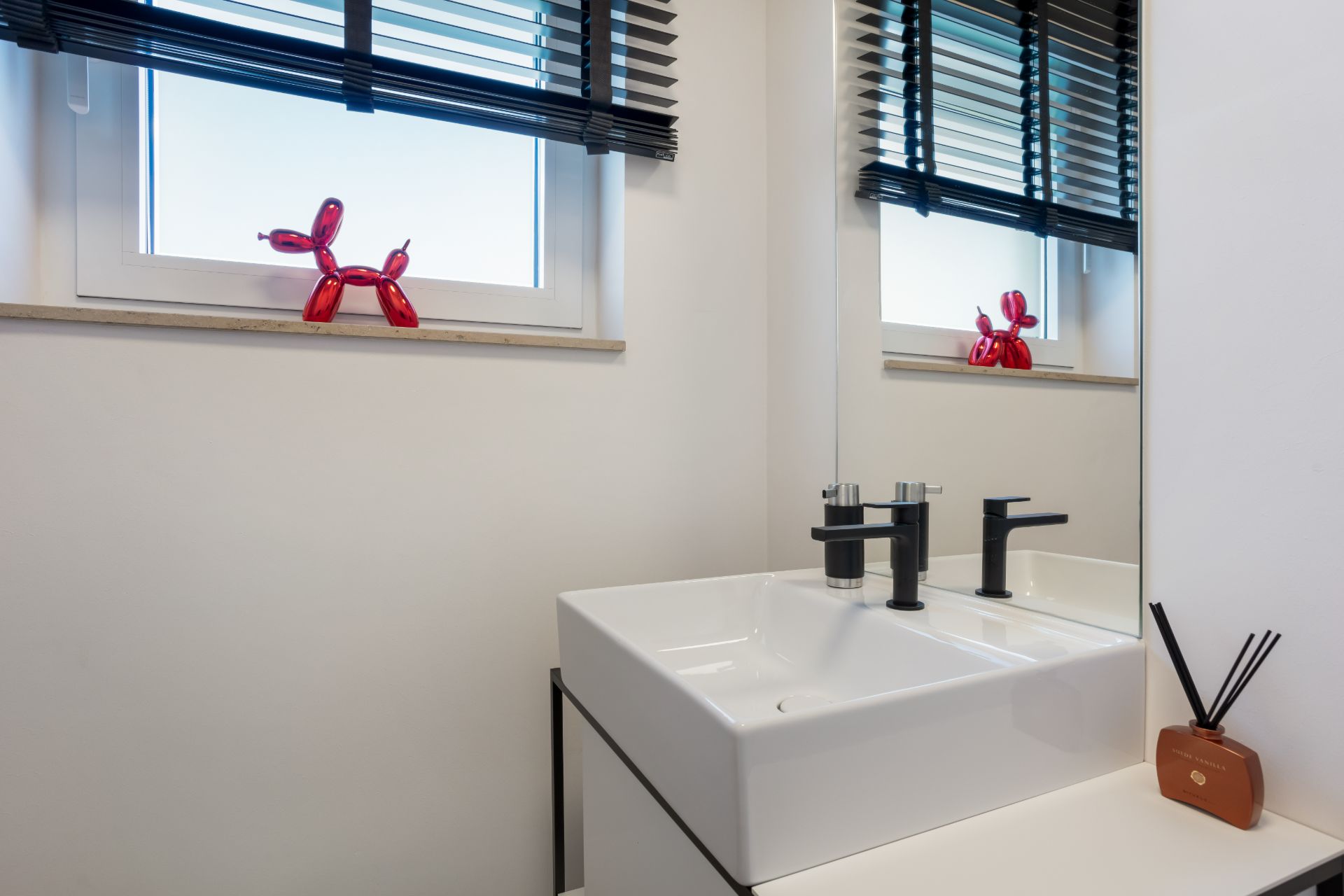 Modernes Badezimmer mit weißem Waschbecken, Fenster mit Jalousien und Deko in Form eines roten Hundes.