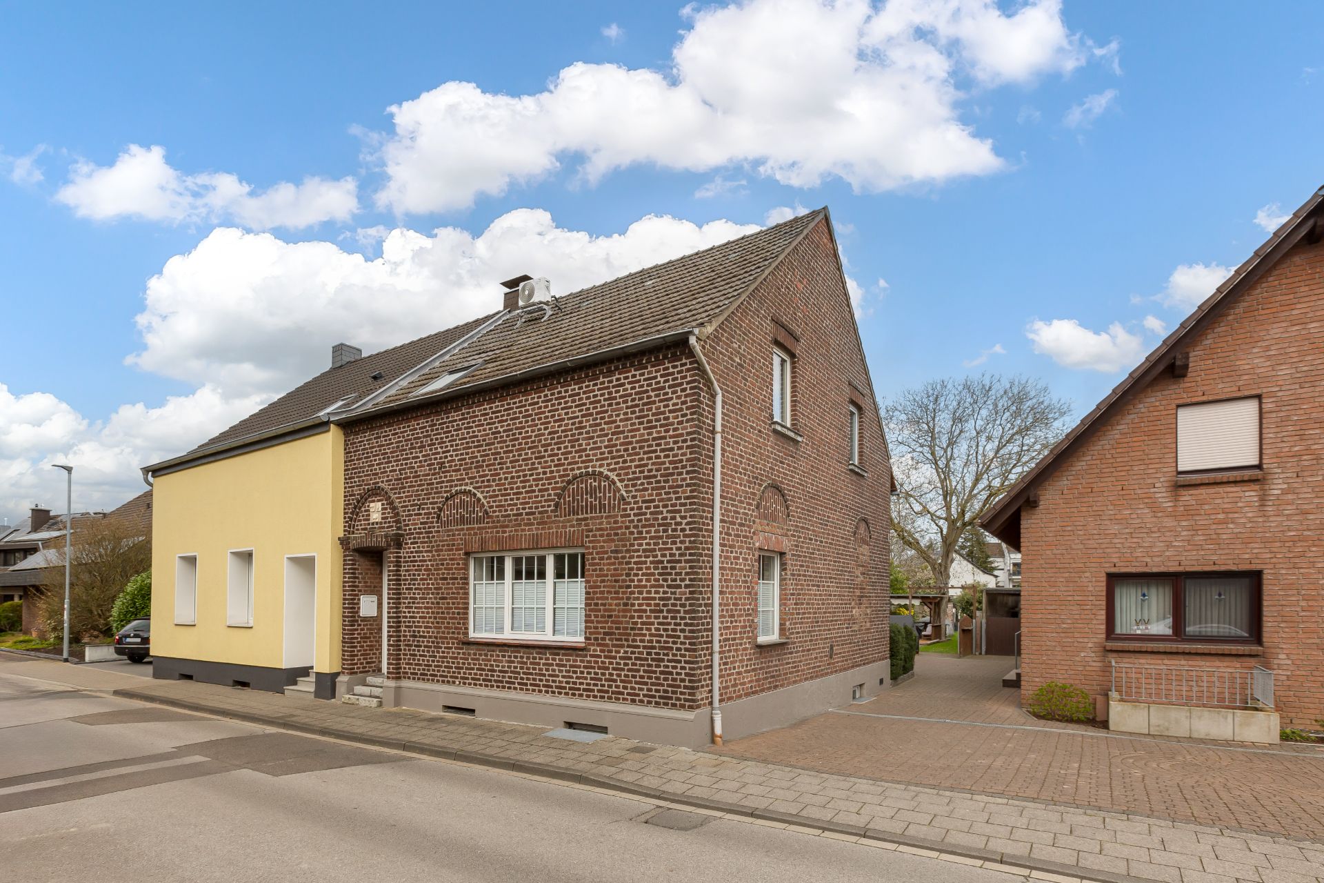 Zweistöckiges Wohnhaus mit brauner Ziegelfassade und gelbem Anbau an einer ruhigen Straße.