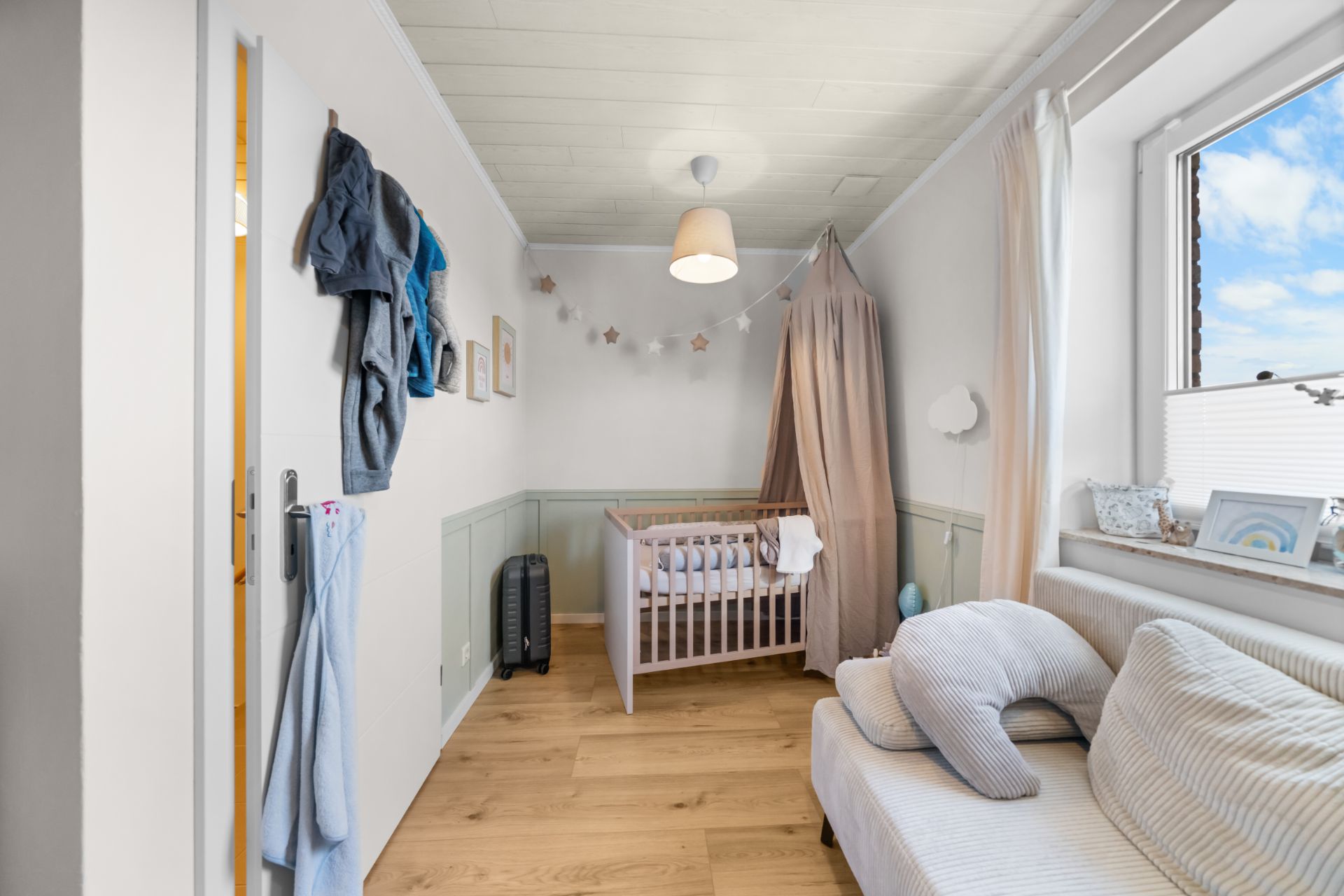 Helles Kinderzimmer mit Babybett, Sofa, Fenster und Holzfußboden.