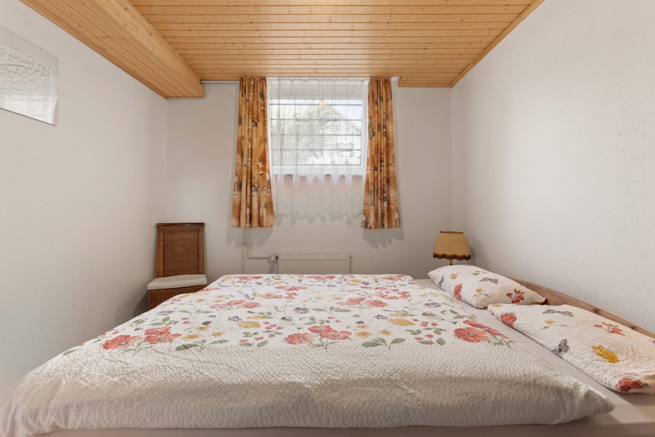 Schlafzimmer mit Doppelbett, geblümter Bettwäsche, Holzdecke und Fenster mit Vorhängen.
