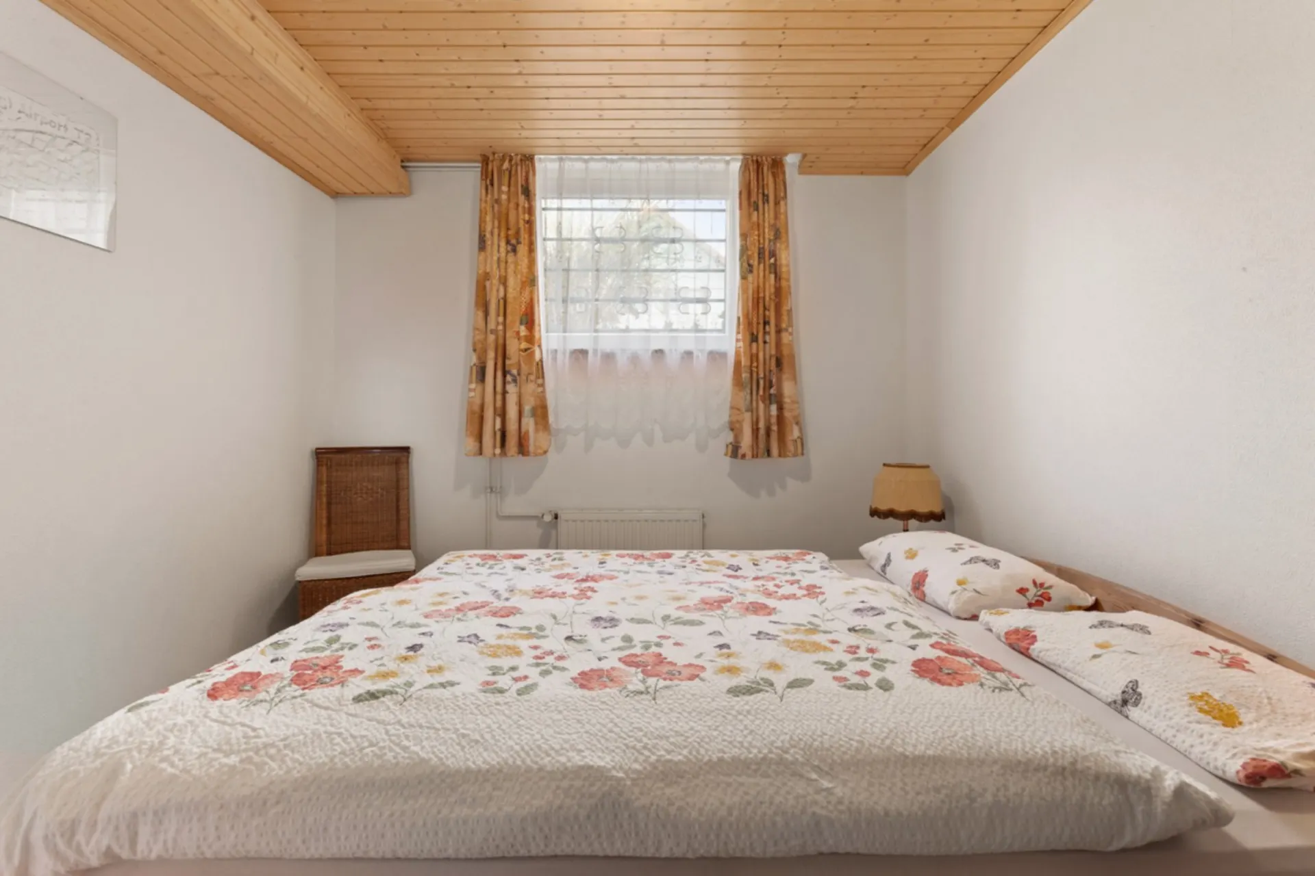 Schlafzimmer mit Doppelbett, geblümter Bettwäsche, Holzdecke und Fenster mit Vorhängen.