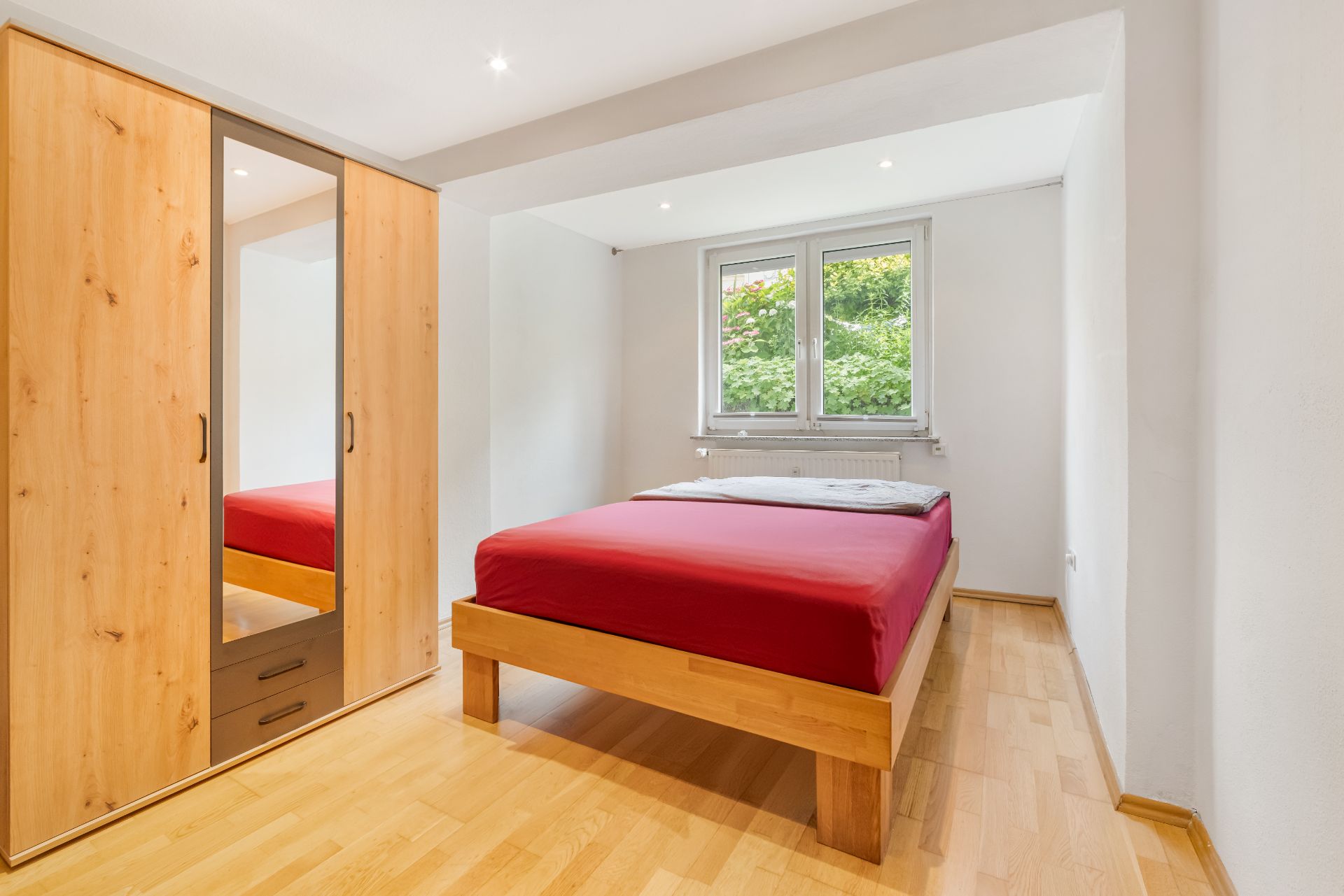 Helles Schlafzimmer mit Holzbett, roter Bettwäsche, Kleiderschrank und Fenster mit Blick ins Grüne.