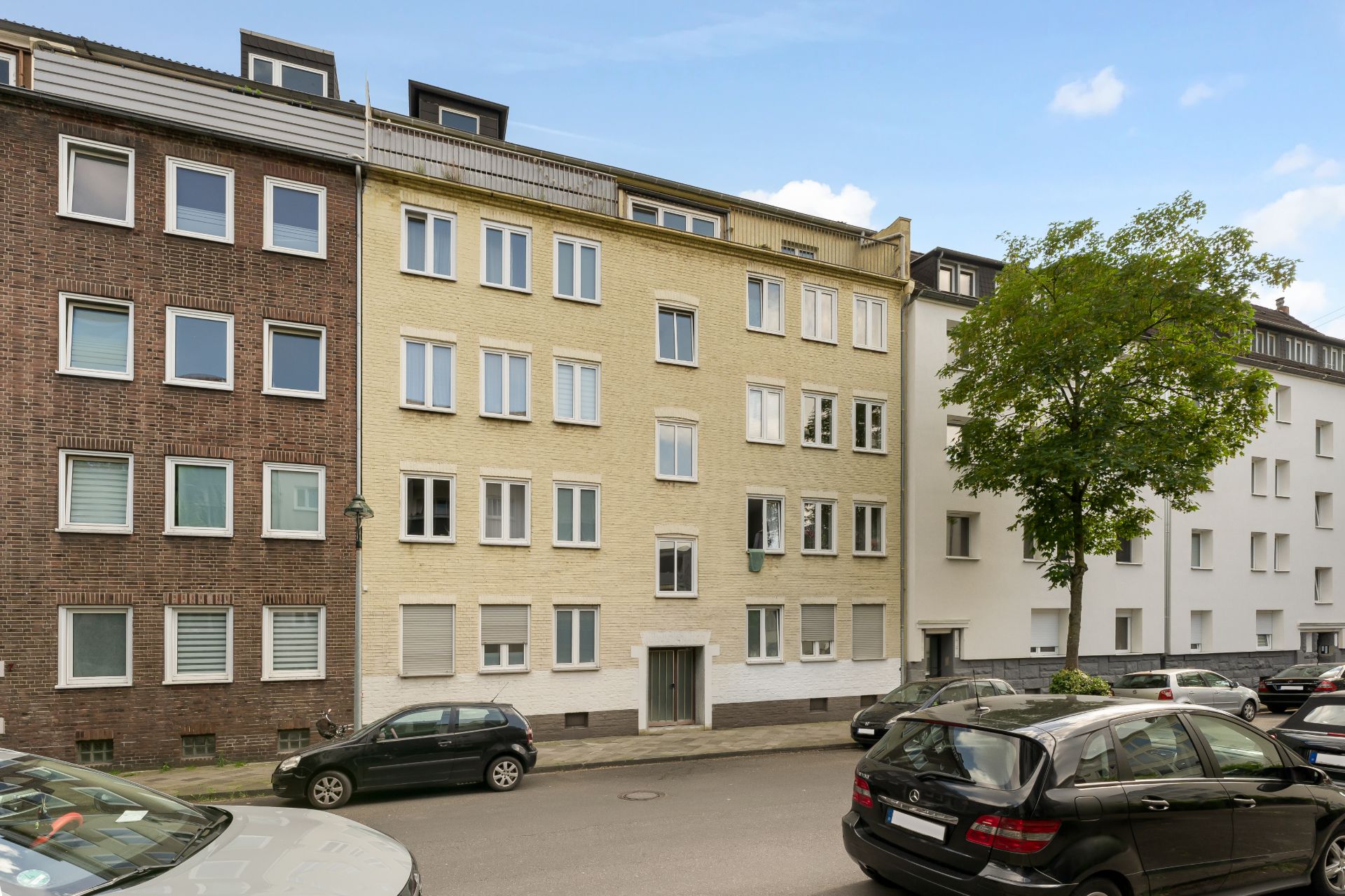Mehrfamilienhaus mit gelber Fassade, mehreren Fenstern und parkenden Autos auf der Straße.