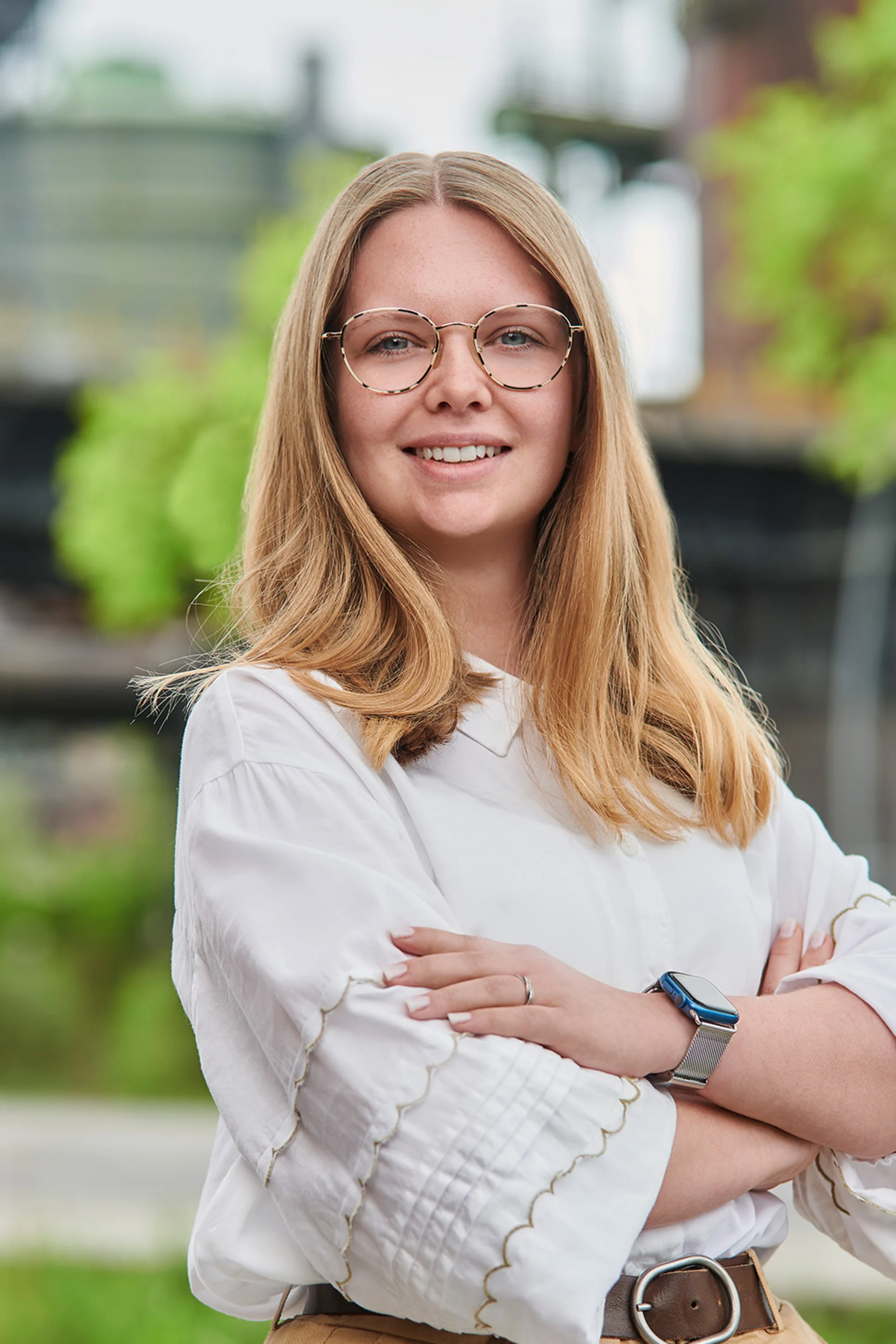 Junge Frau mit Brille und weißer Bluse steht draußen und lächelt, Bäume im Hintergrund.
