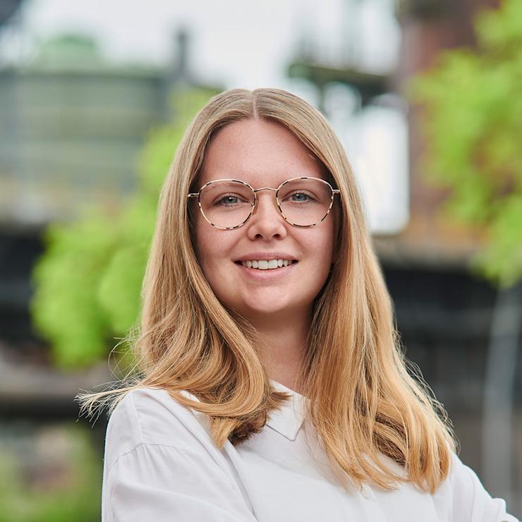 Junge Frau mit Brille und weißer Bluse steht draußen und lächelt, Bäume im Hintergrund.