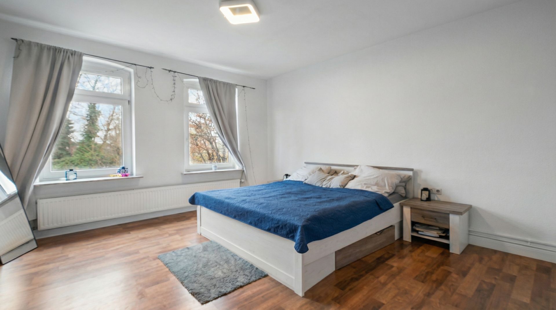 Helles Schlafzimmer mit großem Bett, zwei Fenstern, Vorhängen und Holzboden.