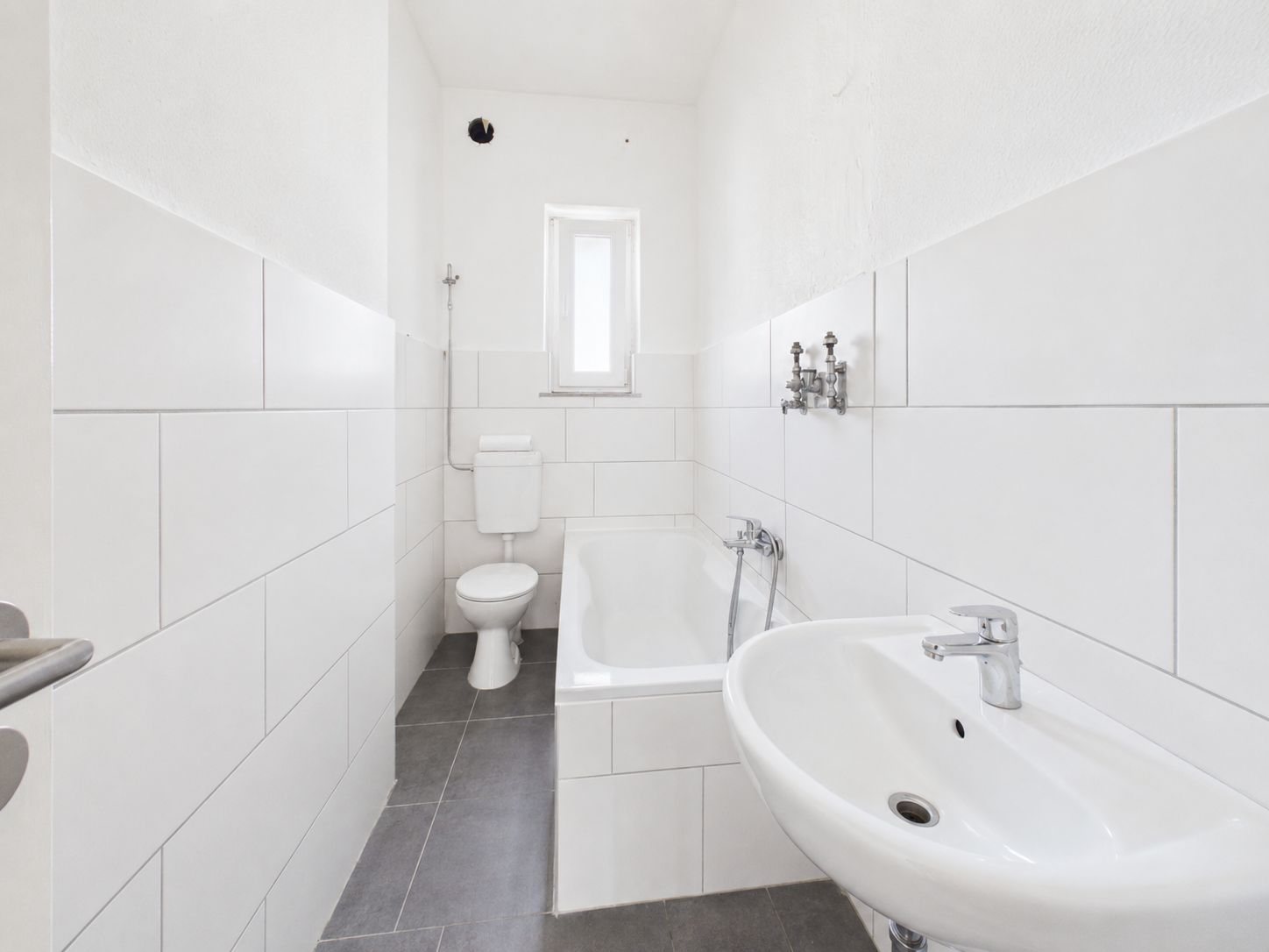 Helles Badezimmer mit weißer Badewanne, Waschbecken, Toilette und kleinem Fenster.
