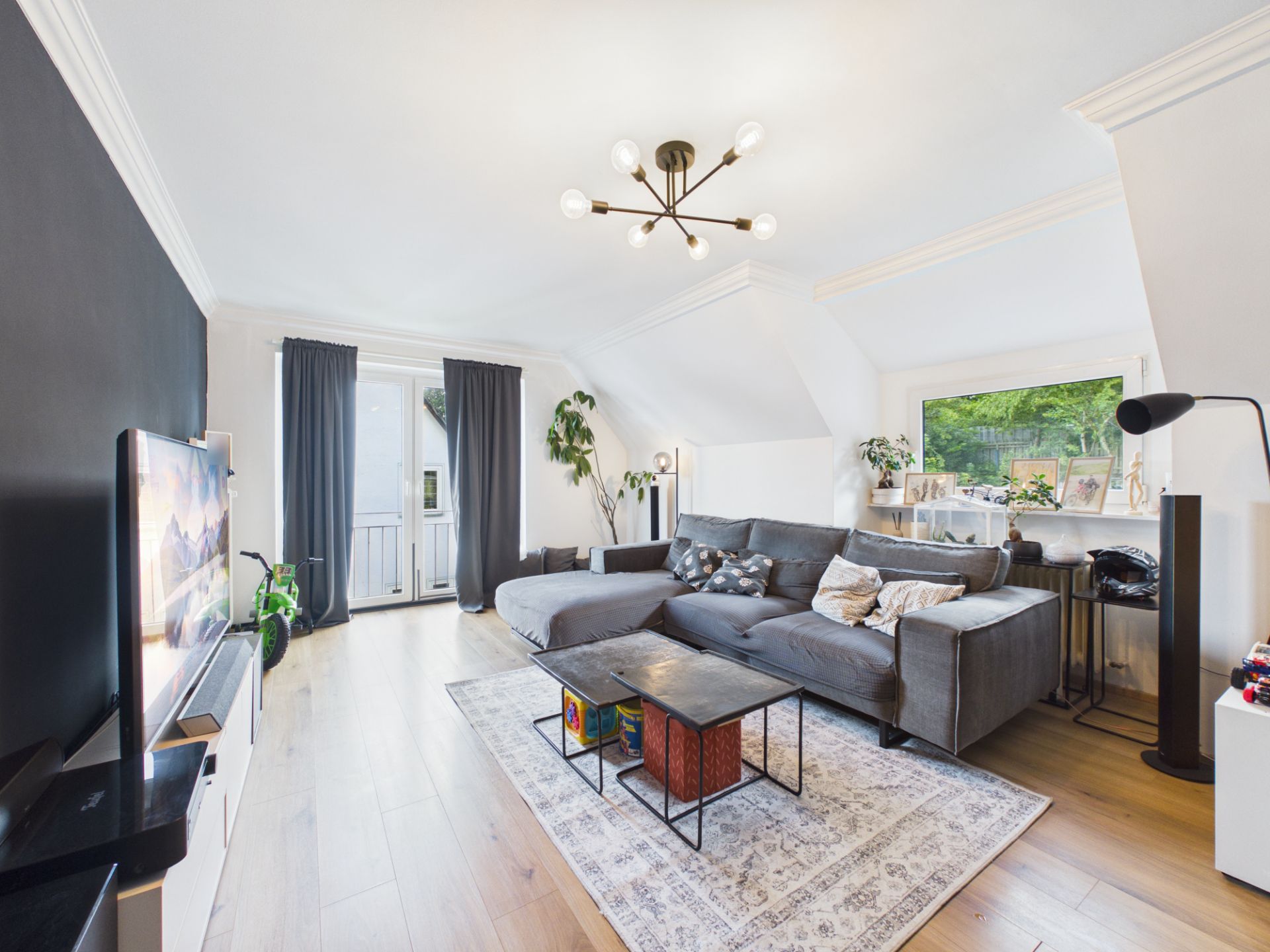 Helles Wohnzimmer mit grauem Sofa, Teppich, Fernseher, Pflanzen und großem Fenster.