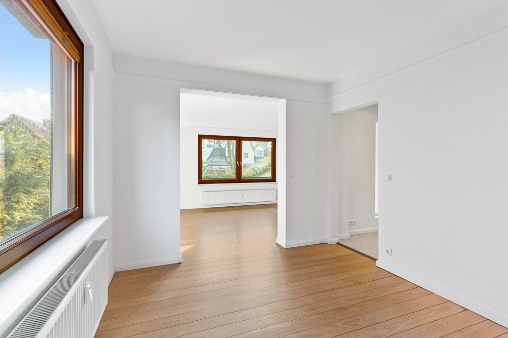Heller Raum mit großen Fenstern, weiß gestrichenen Wänden und Holzfußboden.