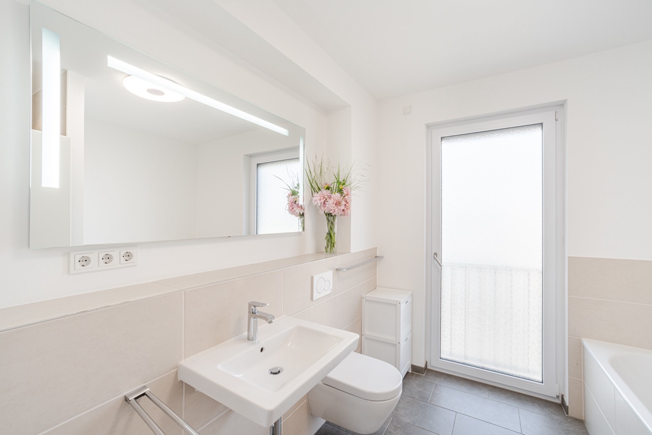 Modernes Badezimmer mit Waschtisch, Spiegel, WC, Fenster und Vase mit Blumen.
