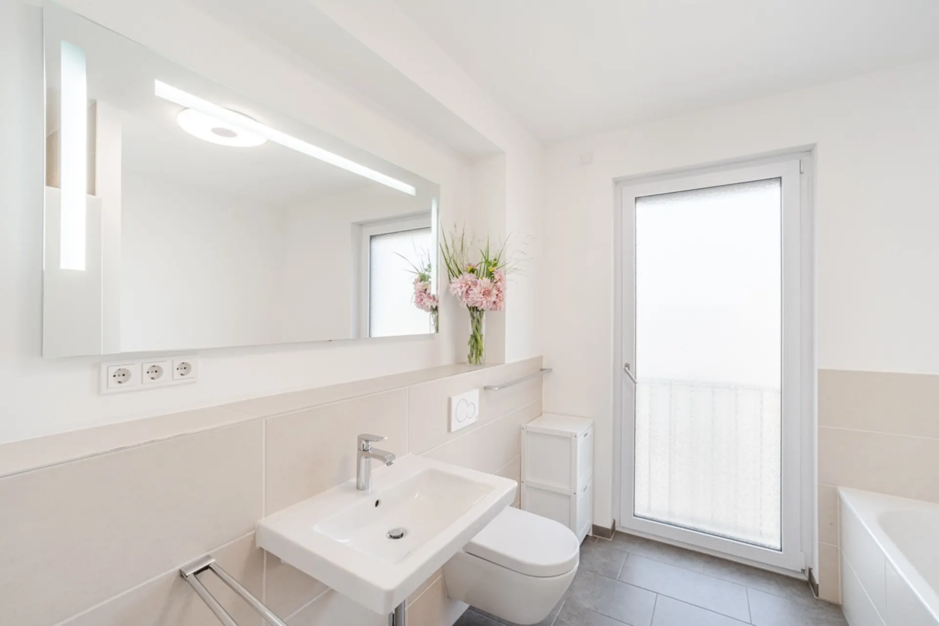 Modernes Badezimmer mit Waschtisch, Spiegel, WC, Fenster und Vase mit Blumen.