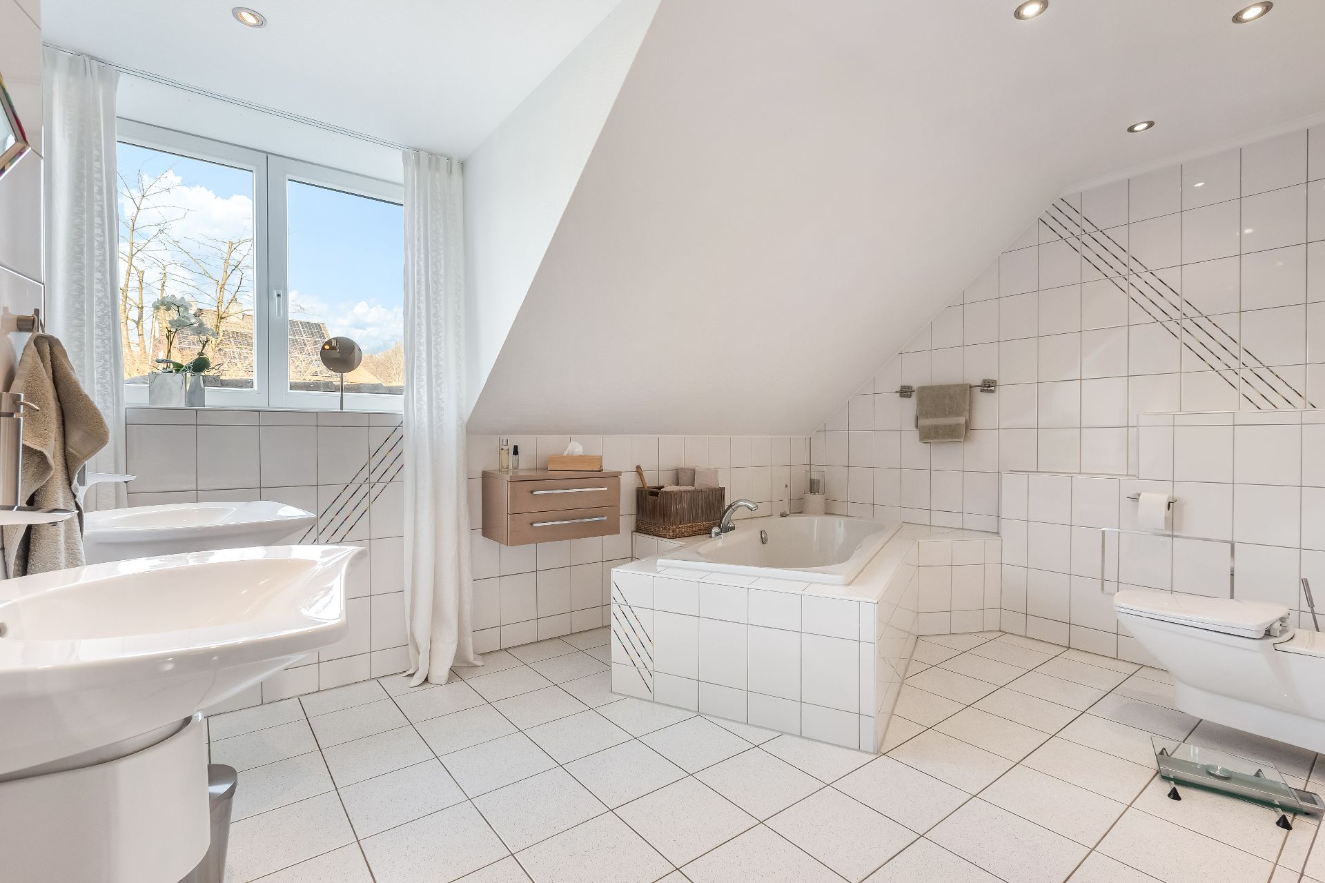 Helles Badezimmer mit Dachschräge, Badewanne, zwei Waschbecken, WC und großem Fenster.